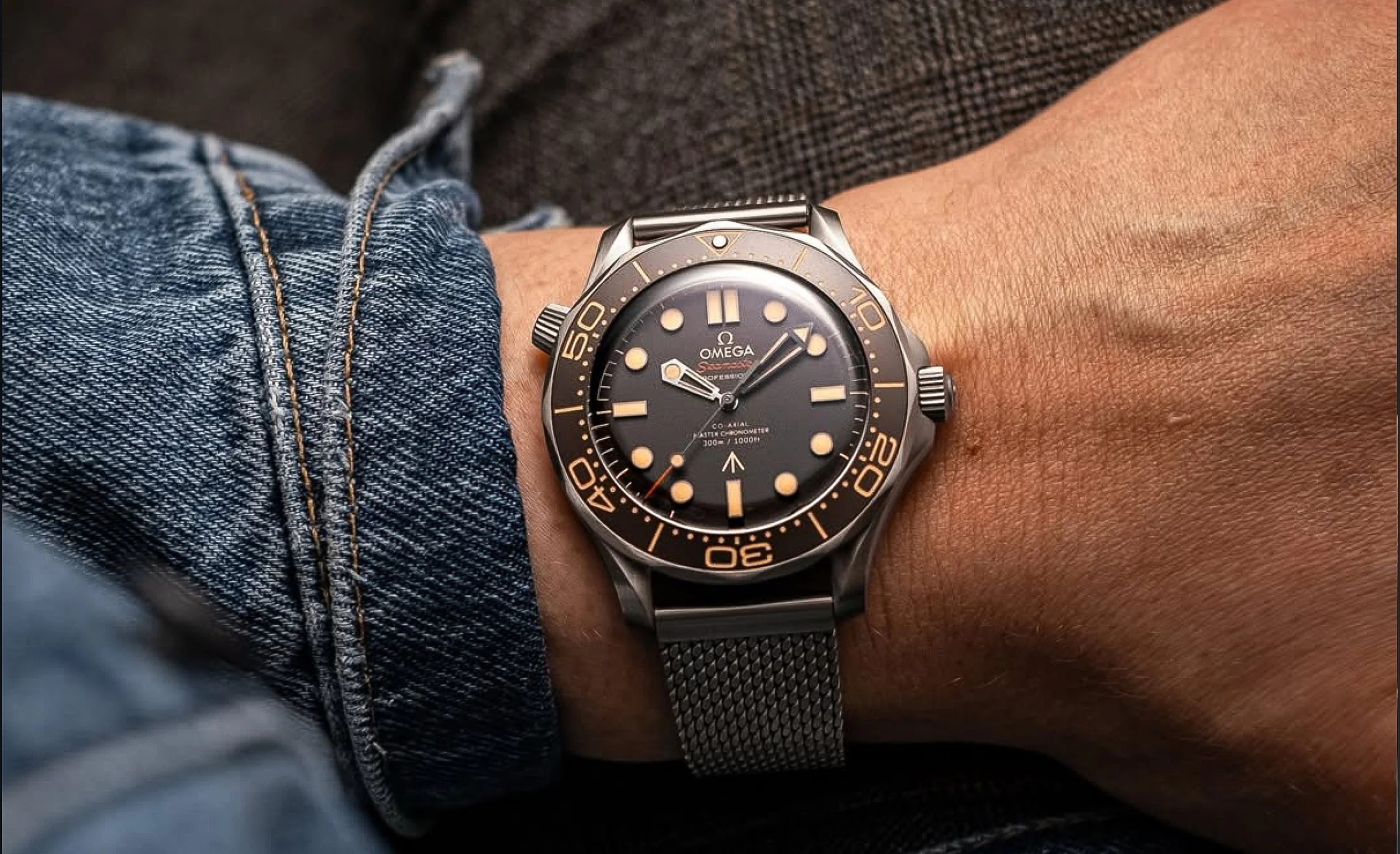 A person's wrist wearing a black Omega Seamaster diver's watch with a stainless steel mesh band, against a background of blue denim and dark fabric.