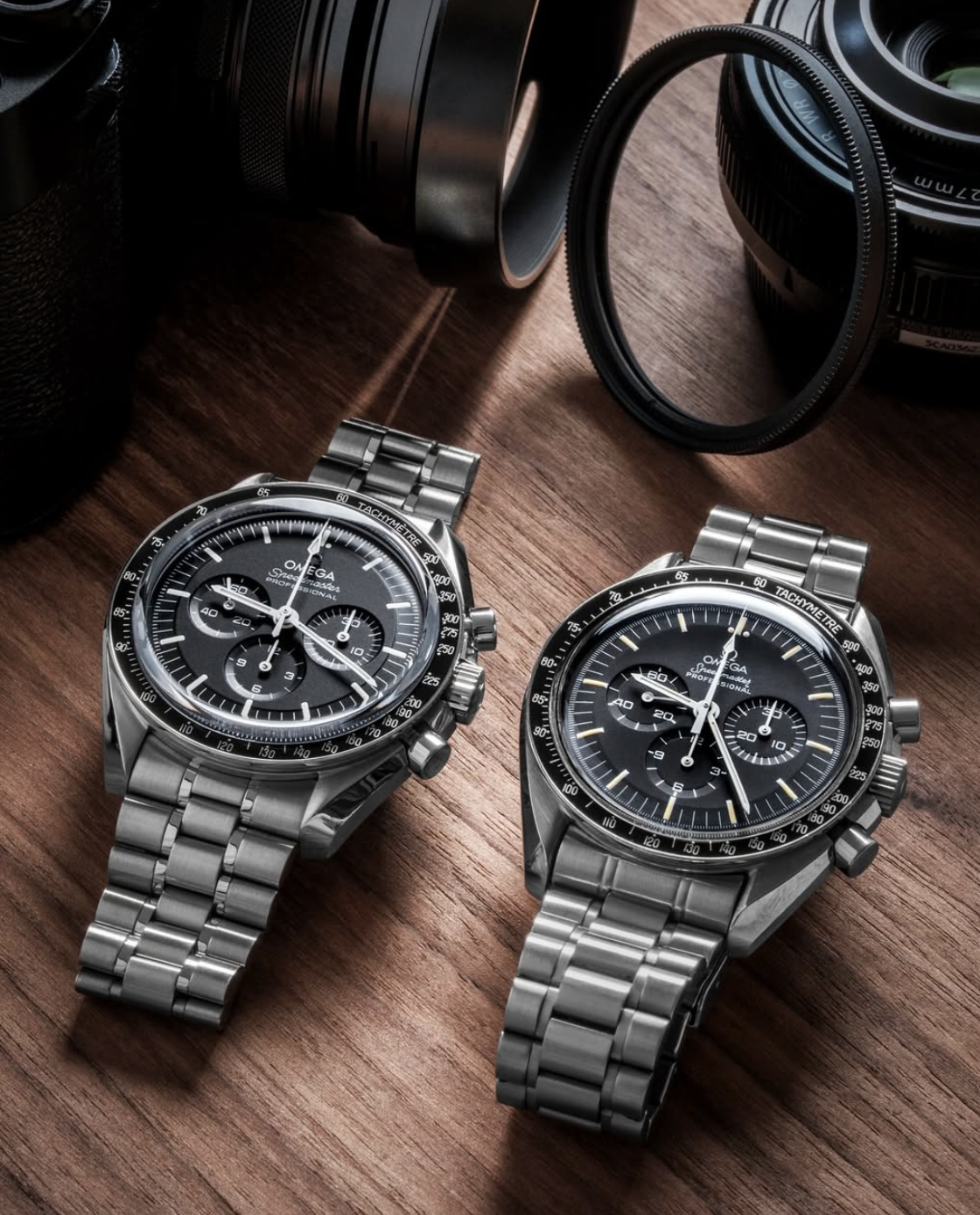 Two Omega Speedmaster watches with metal bands on a wooden surface, with camera lenses and filters in the background.