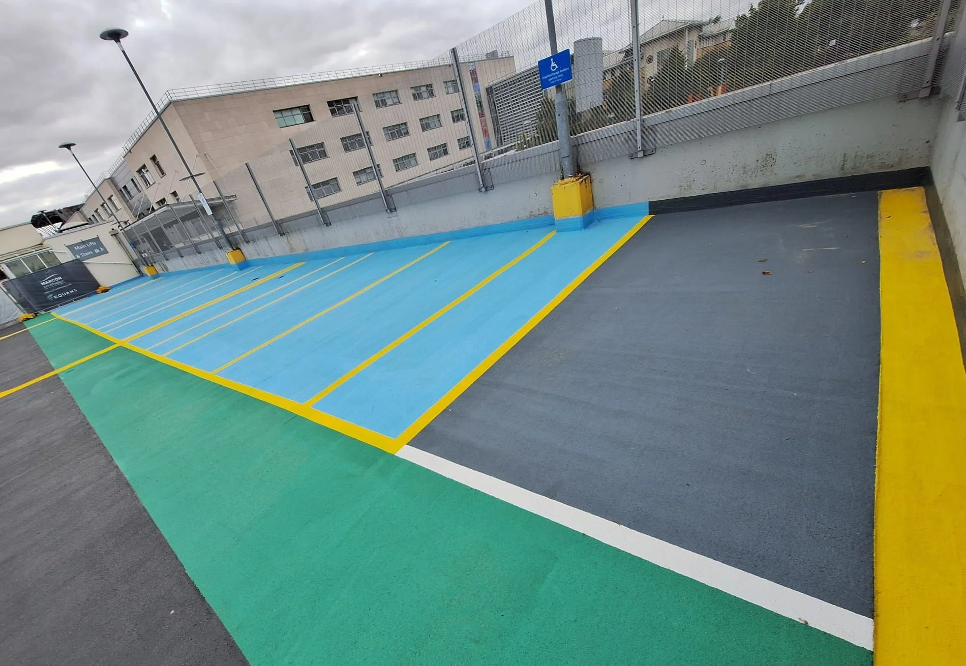 A parking space designated for handicapped parking, painted with blue and yellow lines on an outdoor parking structure with a concrete wall, metal fence, and an informational sign indicating accessible parking.