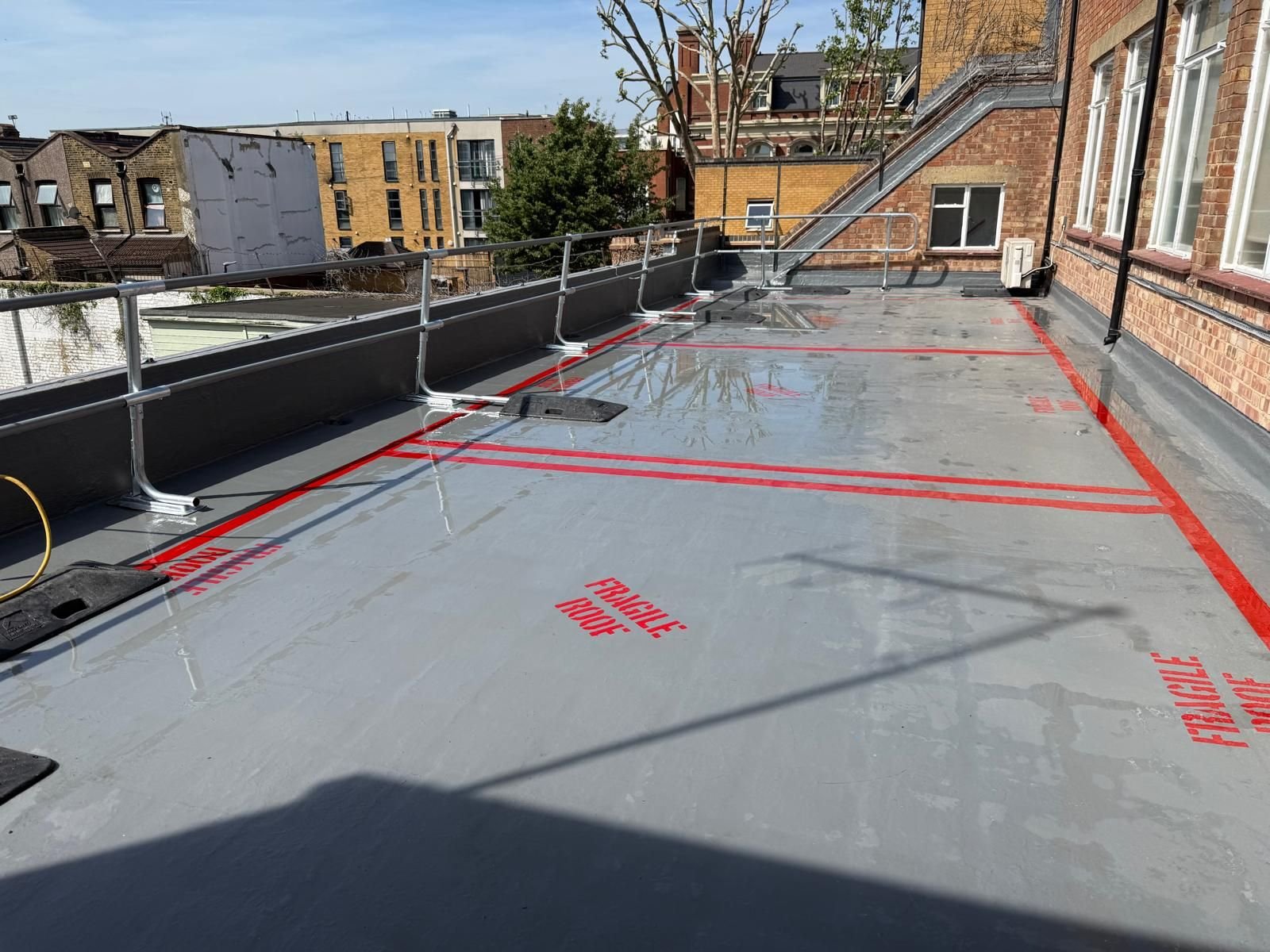 Empty rooftop with waterproofing in progress, marked with red caution tape and protective railings, adjacent to brick building walls and neighboring city buildings.