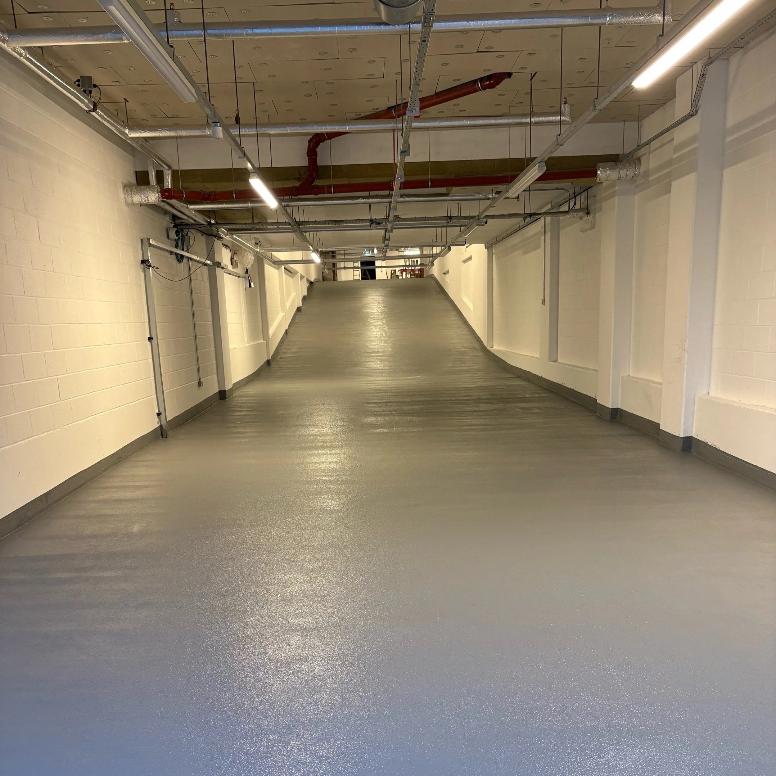 Hallway with white walls, gray flooring, and exposed ductwork and piping along the ceiling. Fluorescent lights are mounted on the ceiling. The hallway has a slight curve and appears to be in an industrial or commercial building.