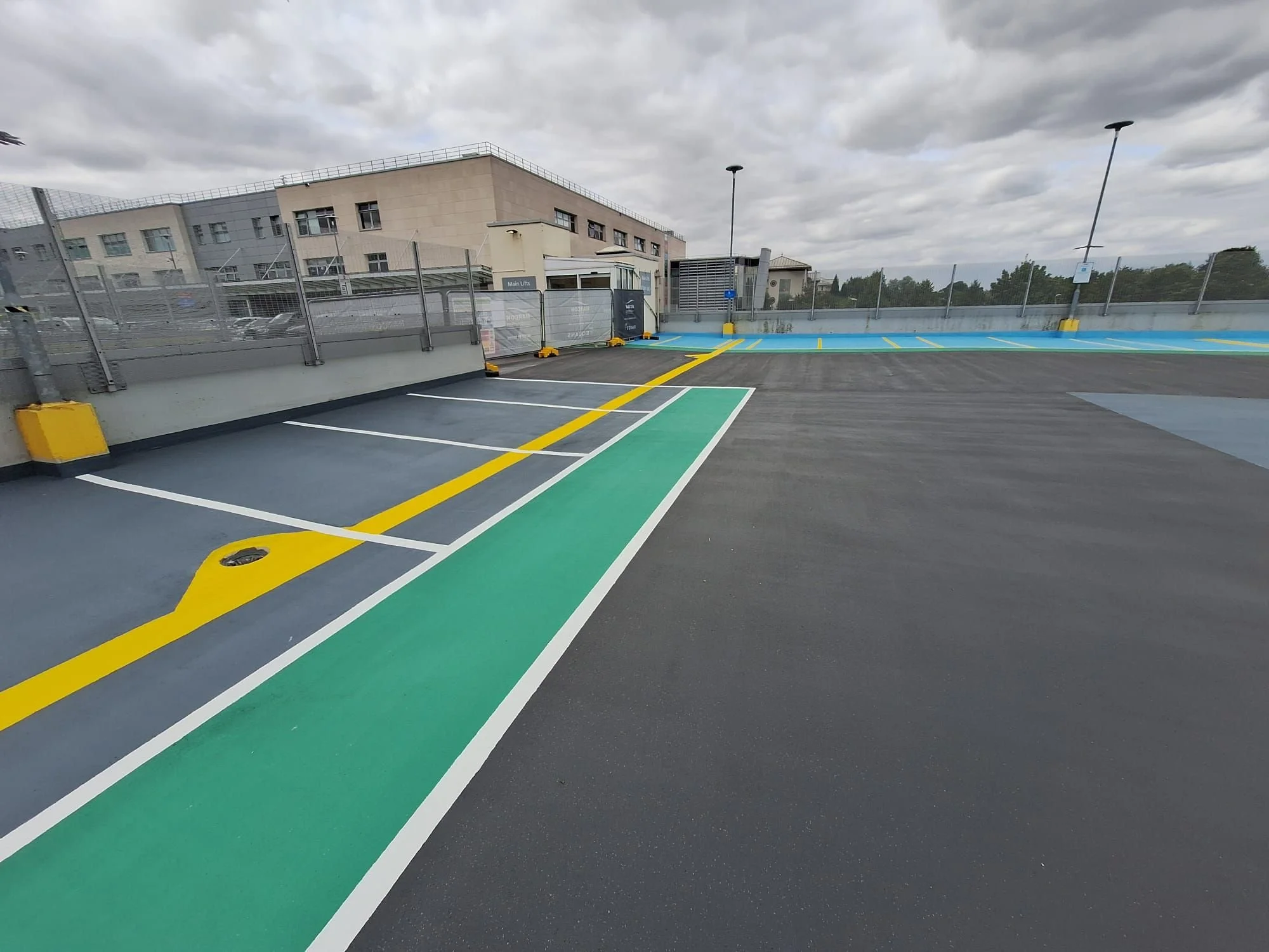 Empty parking lot on a rooftop with green, blue, and black parking spaces, surrounded by a metal fence and some buildings in the background, under a cloudy sky.