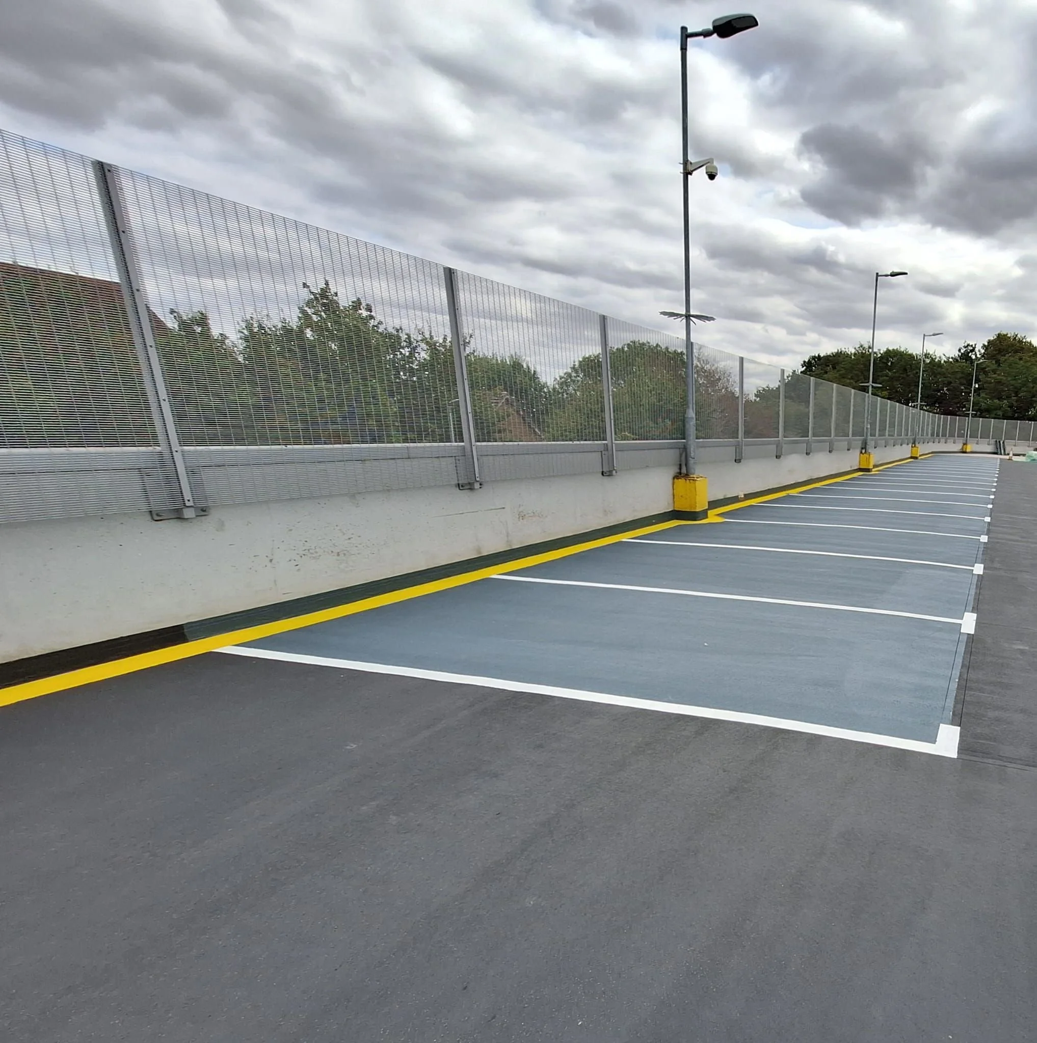 View of an outdoor car park with waterproofed floor and painted yellow and white lines and a metal fence along the edge. There are tall streetlights and cameras mounted on poles. The sky is cloudy with dark gray clouds, indicating overcast weather.