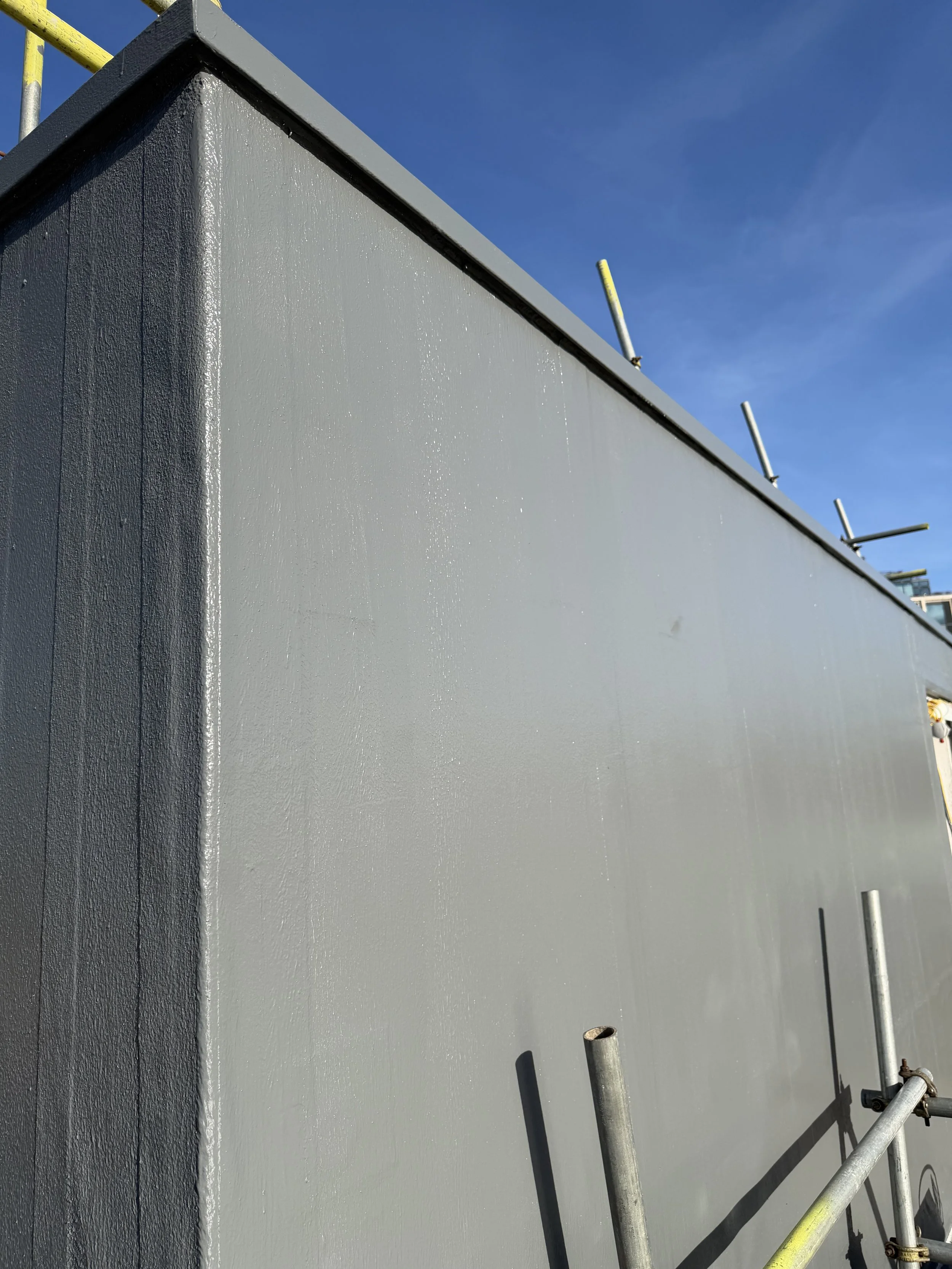 Close-up of a building corner with grey liquid waterproofing, under a clear blue sky, scaffolding poles nearby.