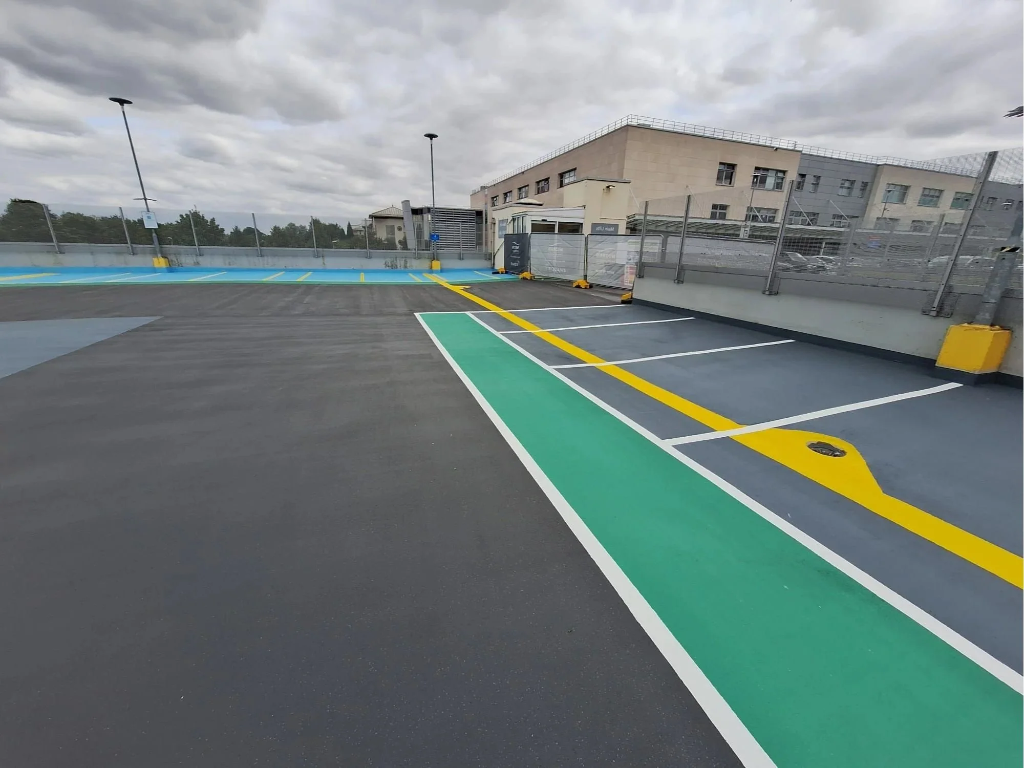 Empty parking lot with green and yellow painted lines, a fence, and a cloudy sky overhead.