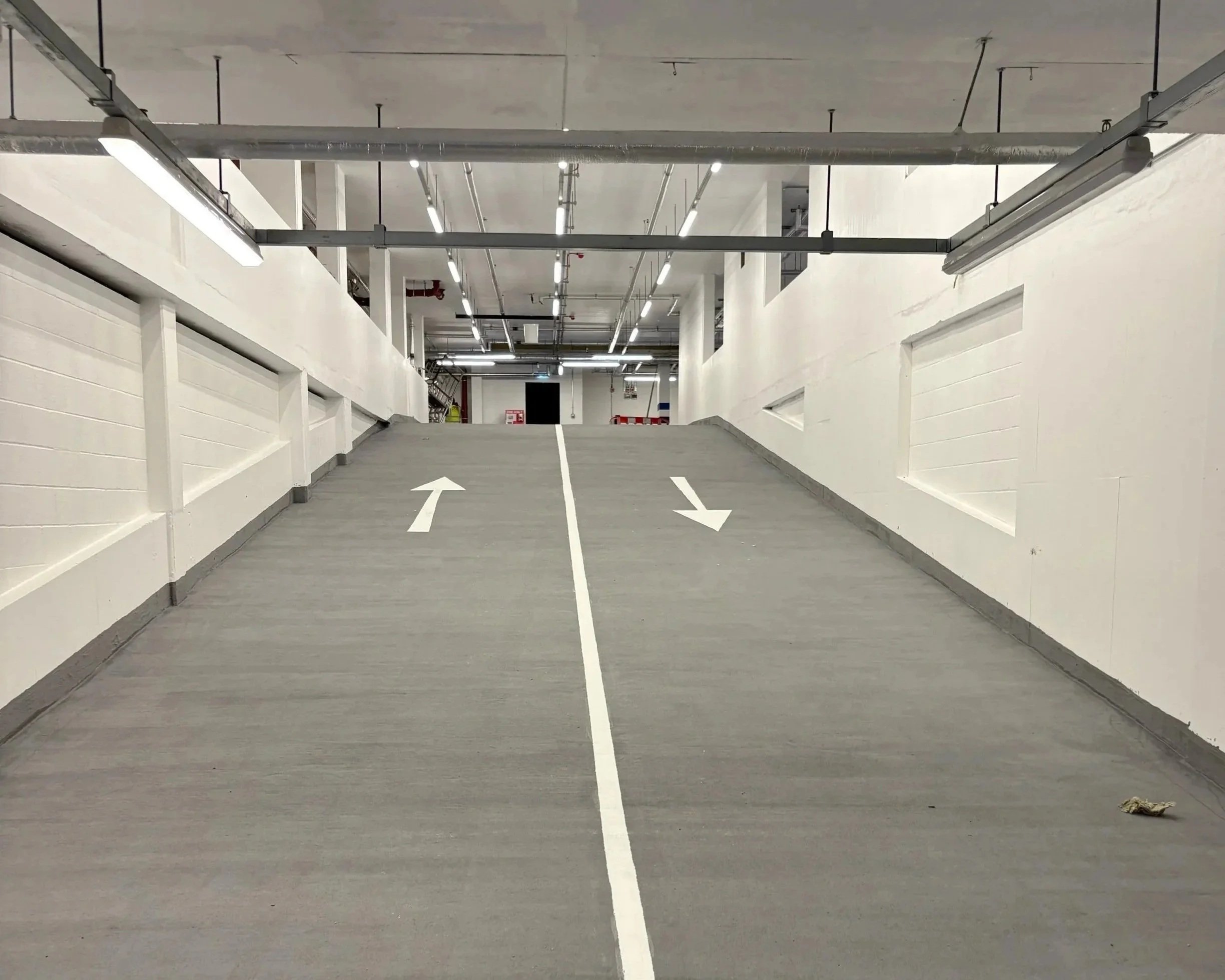 Underground parking garage ramp with arrows pointing in opposite directions for vehicle flow, white walls, ceiling with exposed pipes and fluorescents, and some objects at the top of the ramp.