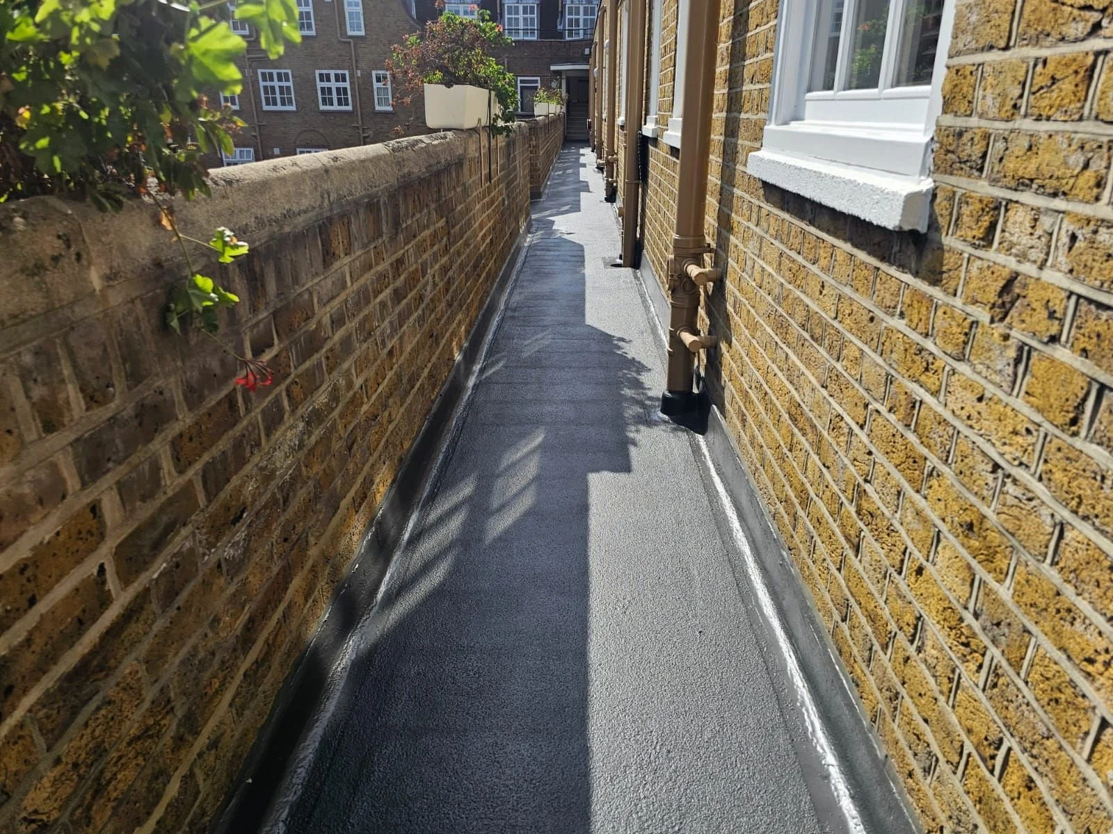 A narrow outdoor walkway between a brick wall on the left and a brick building on the right. The walkway has a freshly laid waterproof membrane, with the right wall featuring a window with white trim and a flower pot with plants. Shadows of a railing
