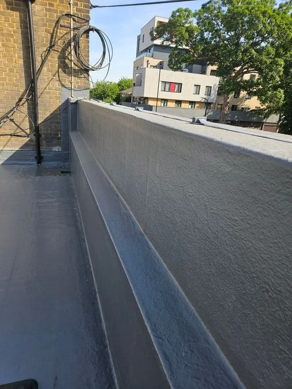 View of a rooftop balcony with a white painted concrete barrier, electrical wires attached to a brick wall on the left, and modern apartment buildings with trees in the background.