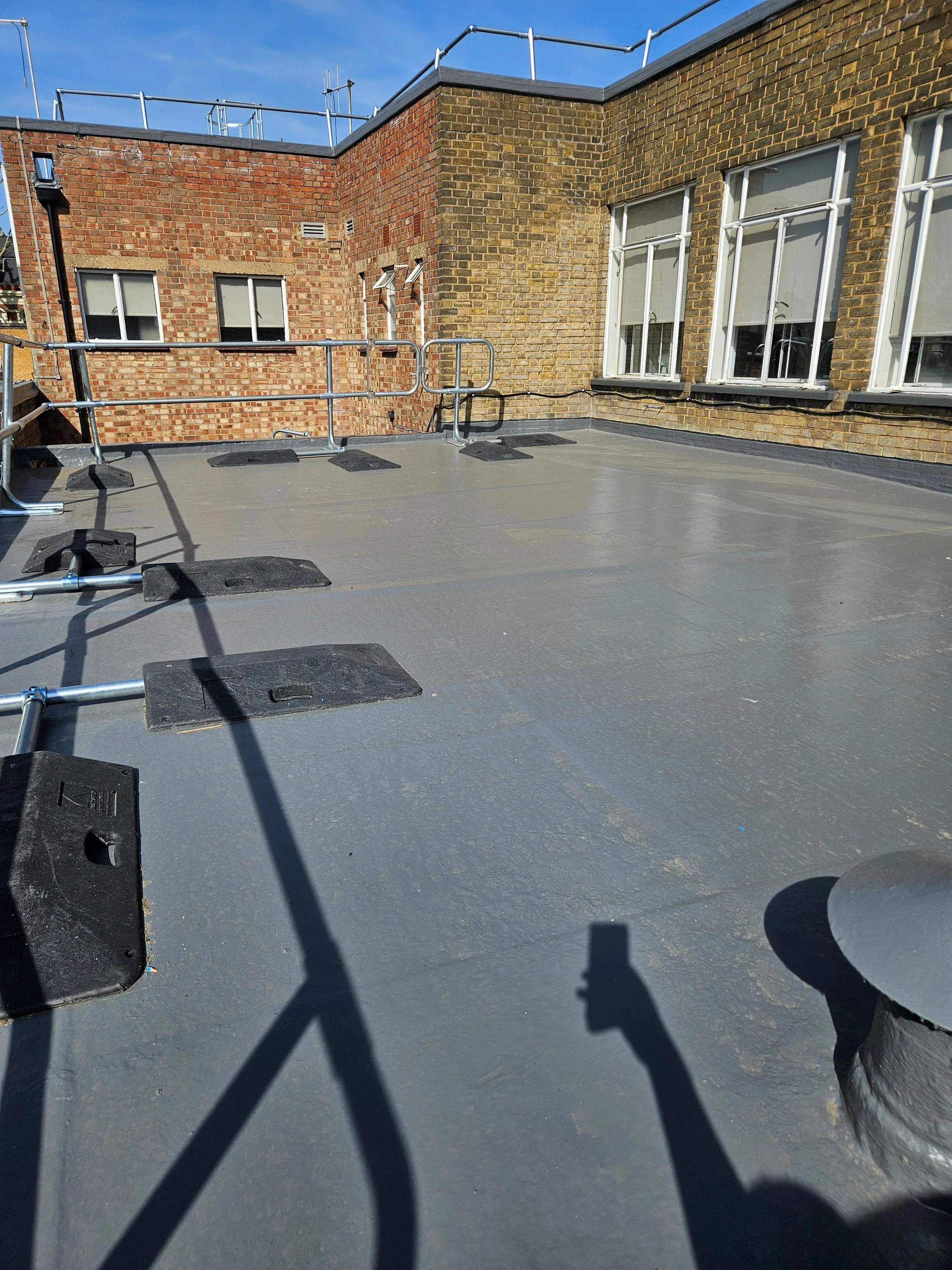 View of a rooftop with fresh waterproofing coating, brick walls, and windows, with scaffolding and blue sky in the background.
