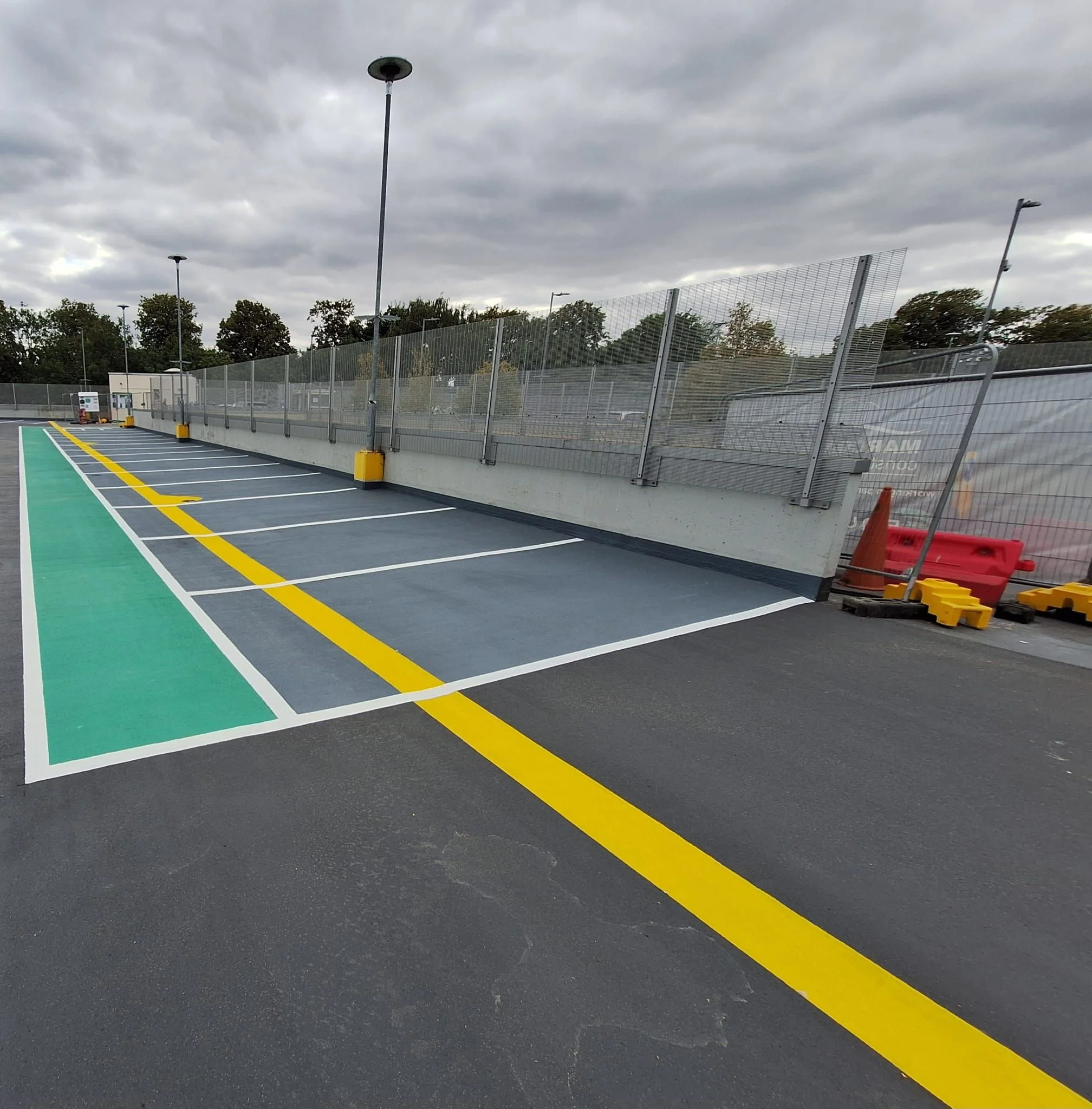 An empty parking lot with designated parking spaces, a tall street lamp, a construction cone, and a fence with a cloudy sky overhead.