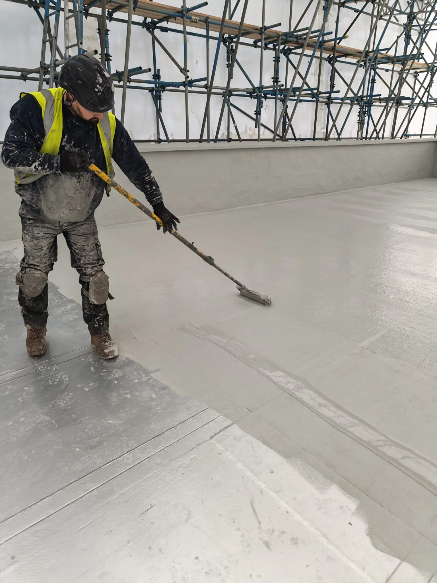 A construction worker wearing a helmet, safety vest, and gloves is smoothing a freshly poured concrete surface with a long-handled tool inside a building under construction. Scaffolding is visible in the background.