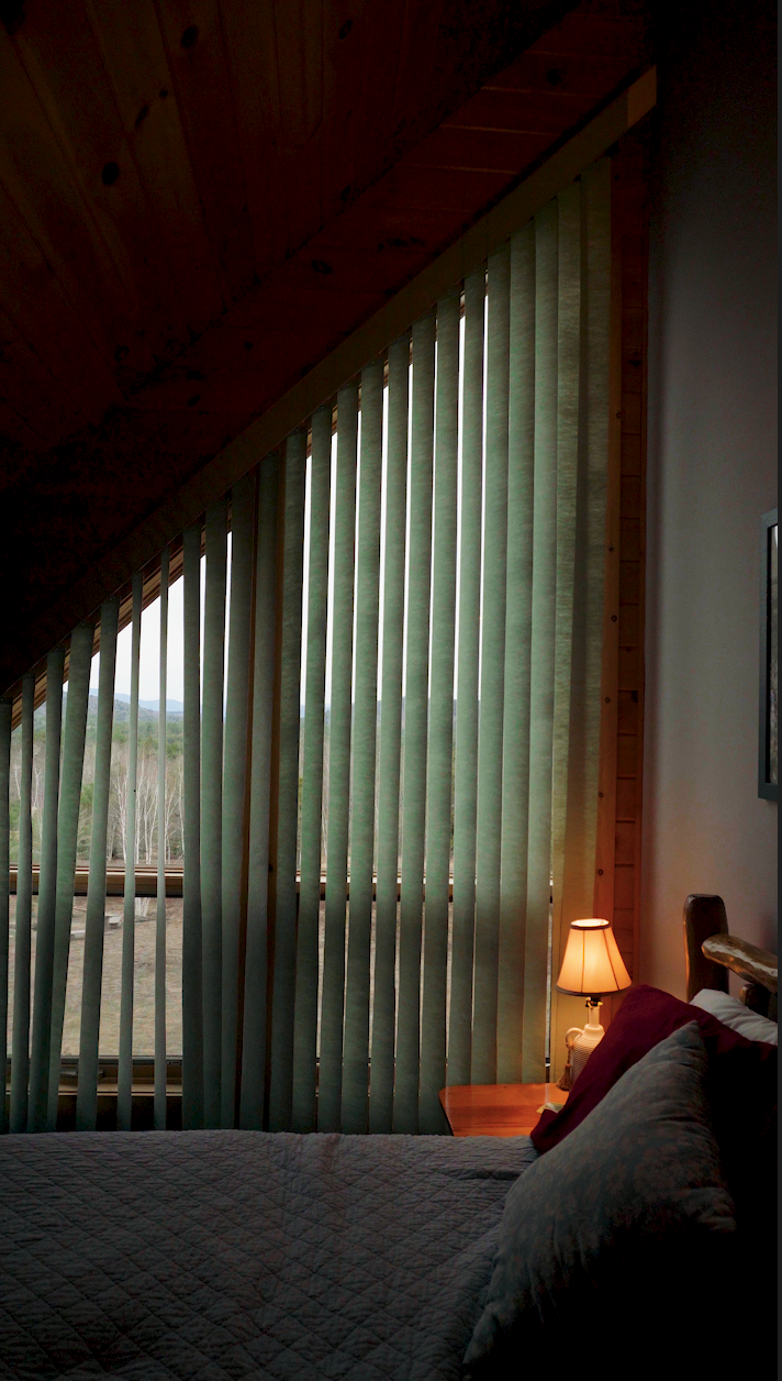 Cozy bedroom with a sloped wooden ceiling, vertical blinds over a large window showing a natural landscape, a bed with pillows and quilt, and a nightstand with a lit bedside lamp.