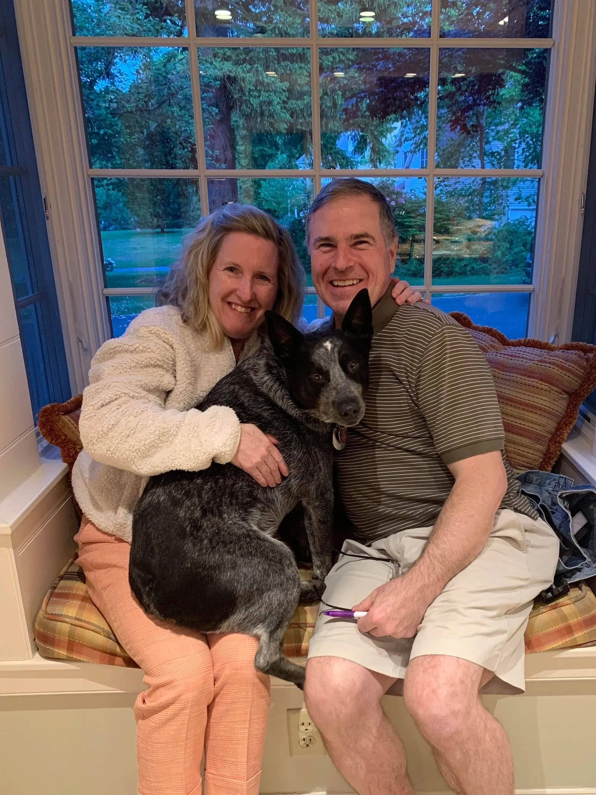 A smiling woman with blonde hair, a smiling man with short hair, and a black and white dog sitting together on a window seat in front of a large window showing trees and a blue sky outside. The woman is wearing a white fuzzy sweater and peach-colored pants, and the man is wearing a striped shirt and khaki shorts. The dog is sitting between them, looking directly at the camera.