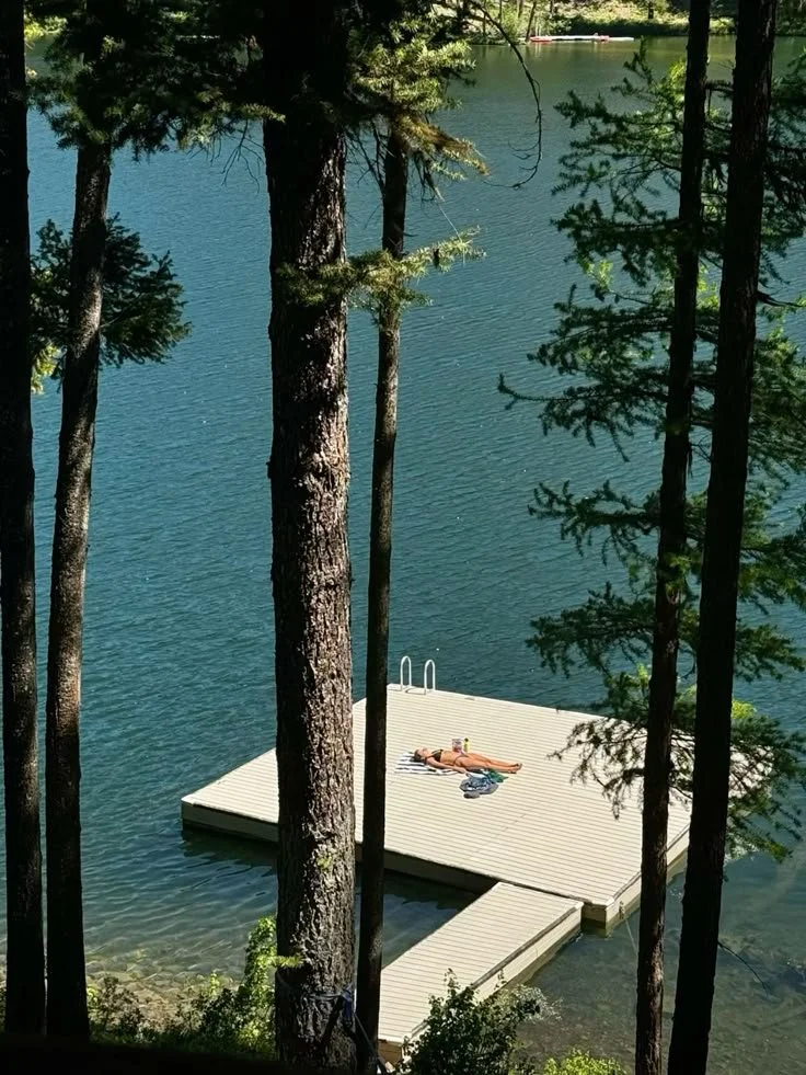 Person sunbathing on a dock by a lake, surrounded by pine trees.