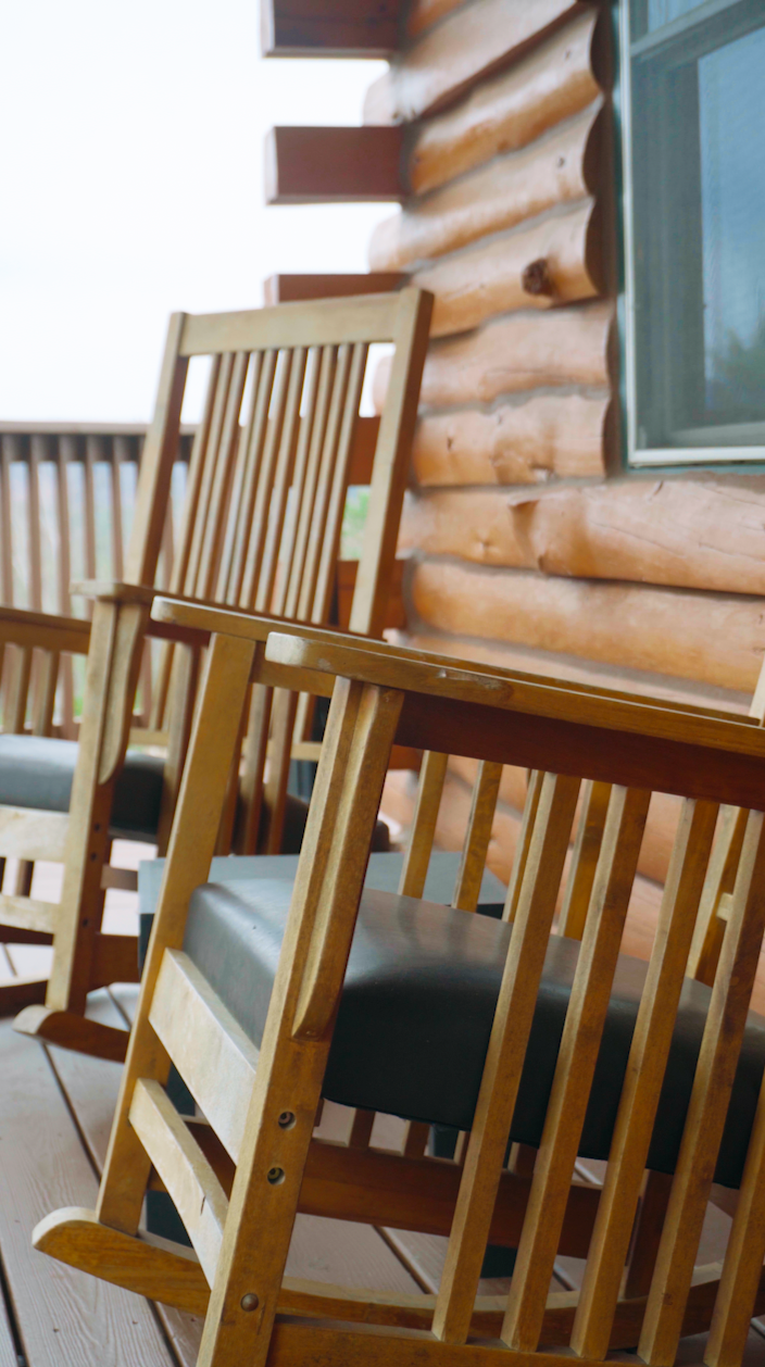 Exterior view of wooden rocking chairs on a porch with a log cabin wall and window.