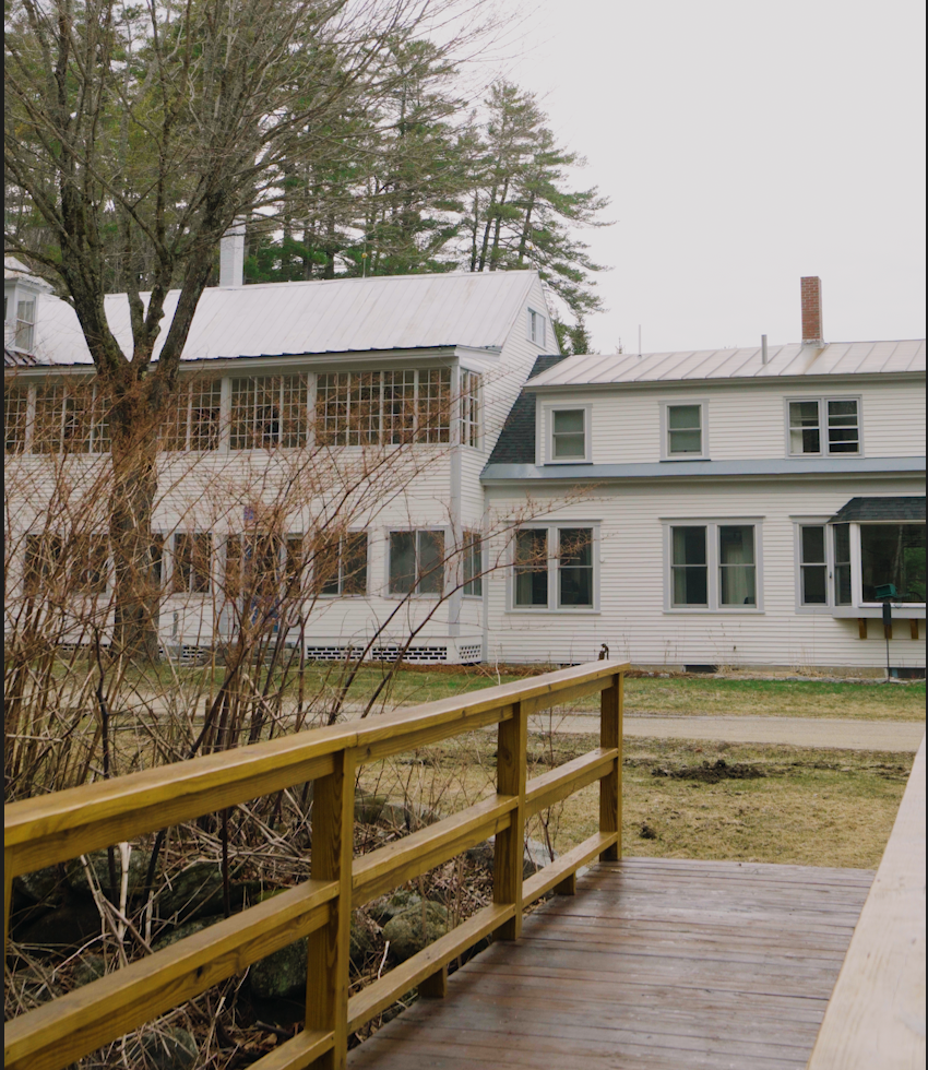 A view from a wooden porch showing a white house with multiple windows, a metal roof, and a chimney, surrounded by leafless trees and a grassy yard.