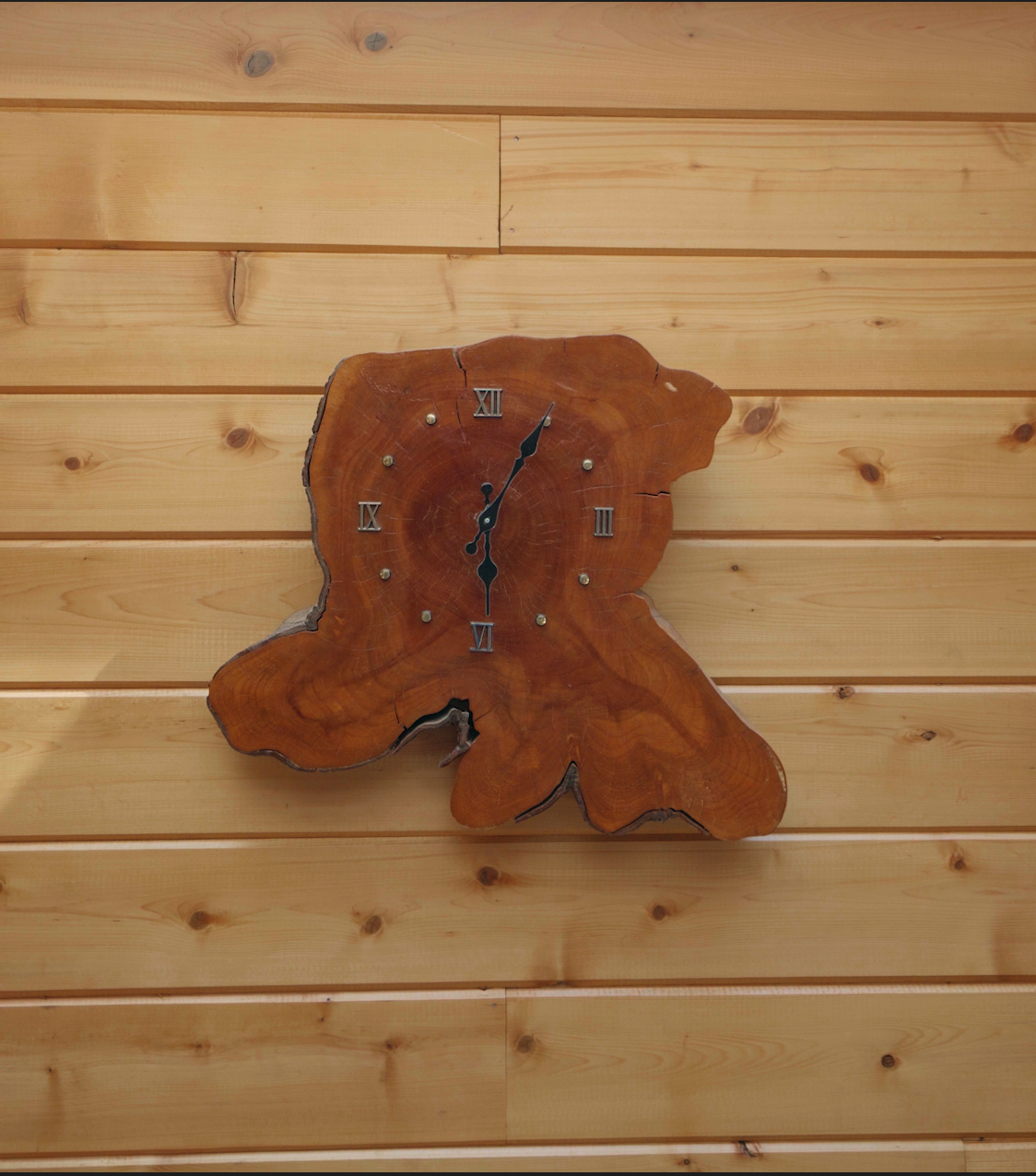 A wall clock mounted on a wooden wall, with a clock face made from a piece of natural wood with irregular edges and a crack, featuring Roman numerals and black clock hands showing the time as 12:05.