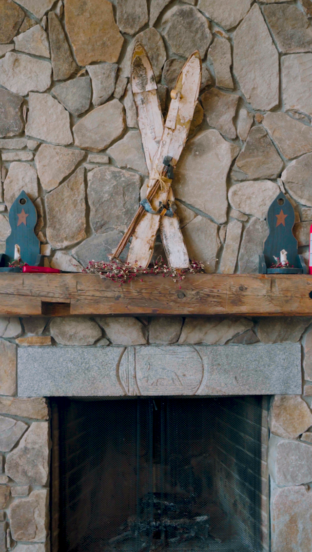 A stone fireplace with a rustic wooden mantel decorated with two blue wooden candle holders, a bundle of dried flowers, and a vintage pair of wooden skis with ski poles leaning against the stone wall.