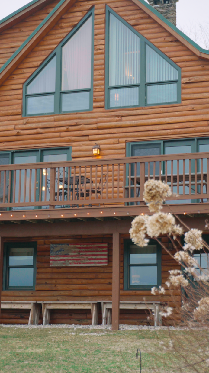 A multi-story wooden house with large triangular window panels on the upper floor, a balcony with a wooden railing, and an American flag mural on the exterior wall.