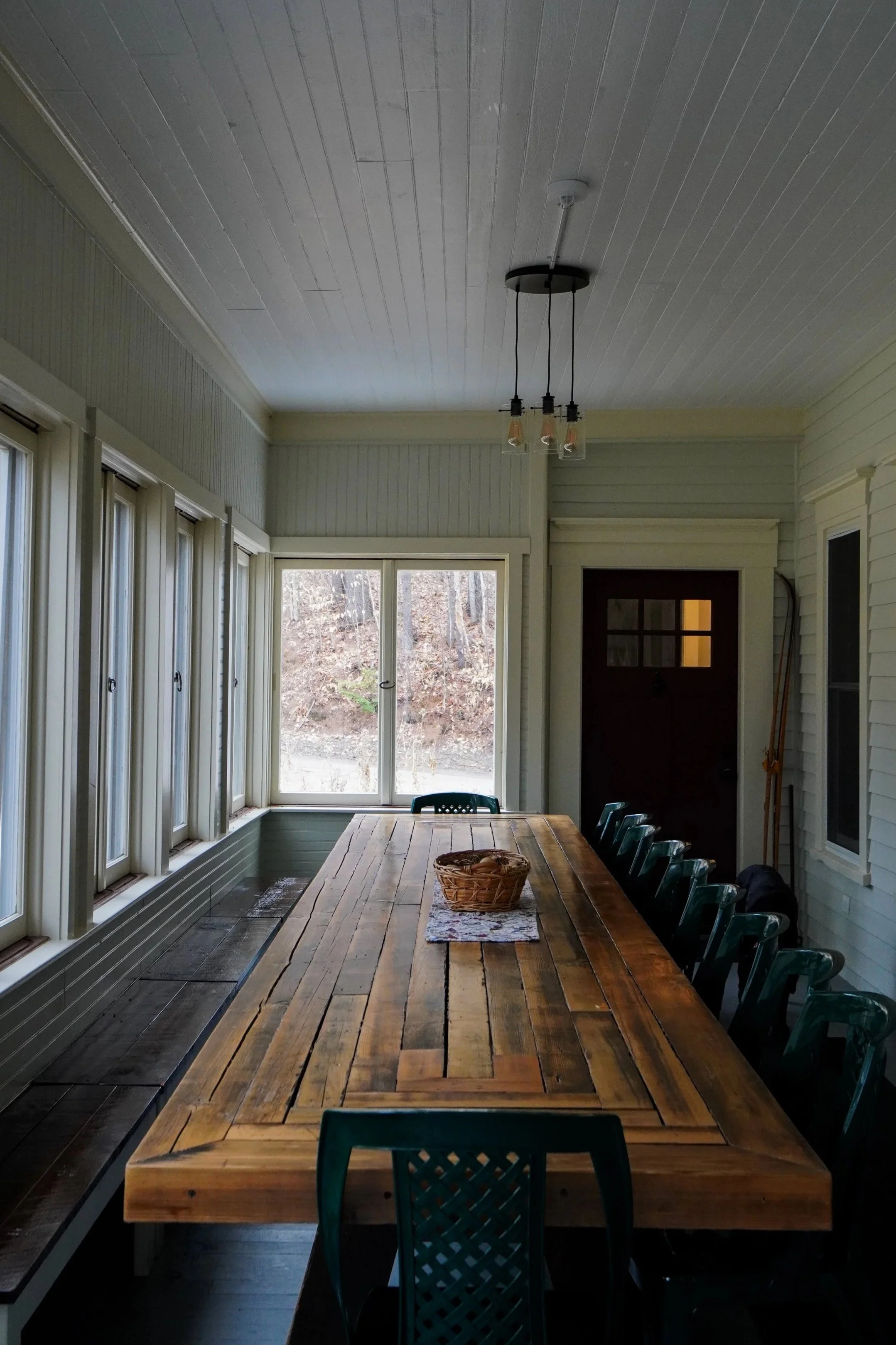 A room with a long wooden dining table surrounded by green plastic chairs. There is a basket and a cloth on the table. The room has window shutters on the left, large windows in the center, and a door at the back. A ceiling fixture with three hanging lights is above the table.