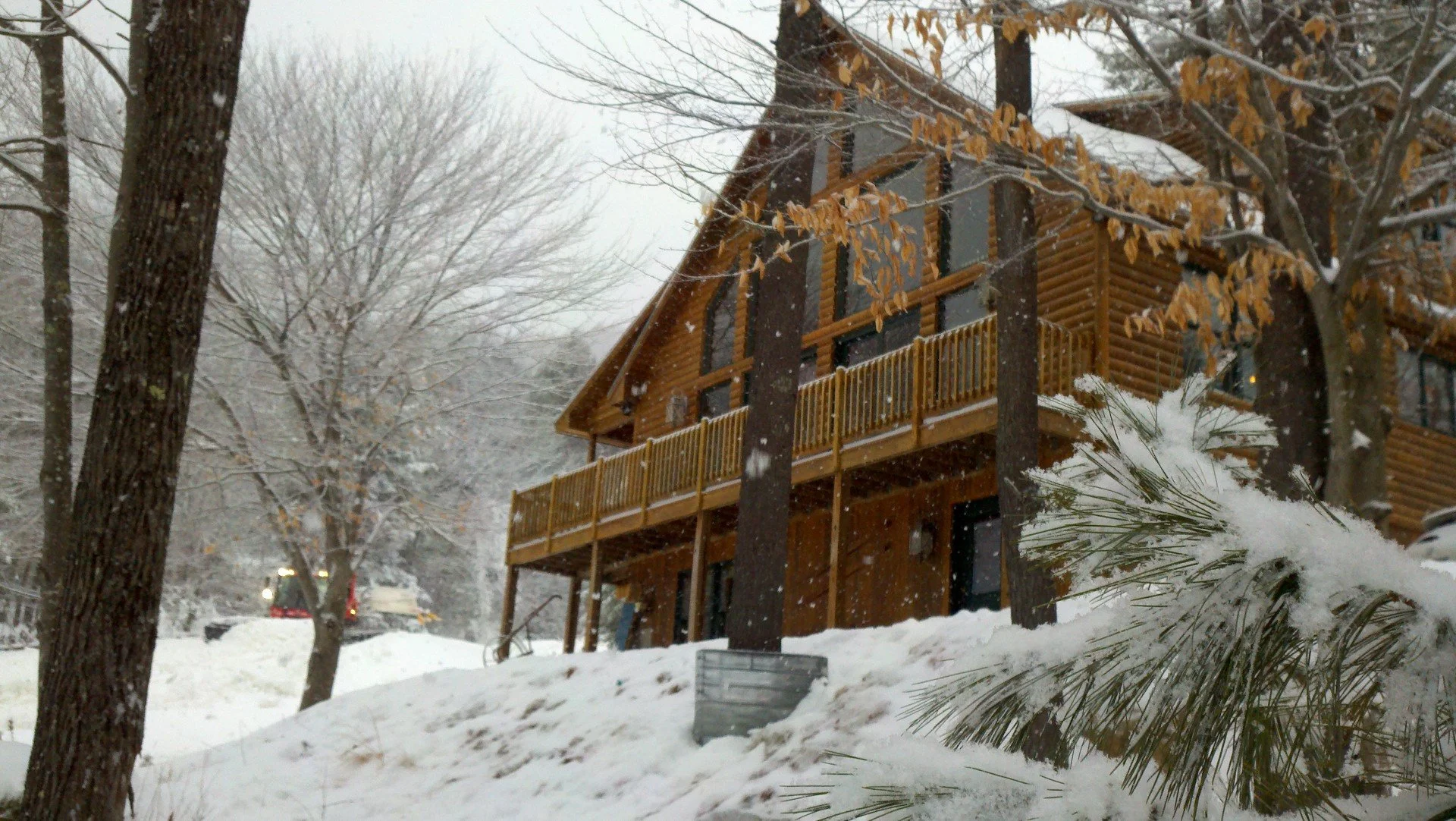 A wooden house with a balcony in a snowy landscape, surrounded by trees with some leaves and snow on the branches.