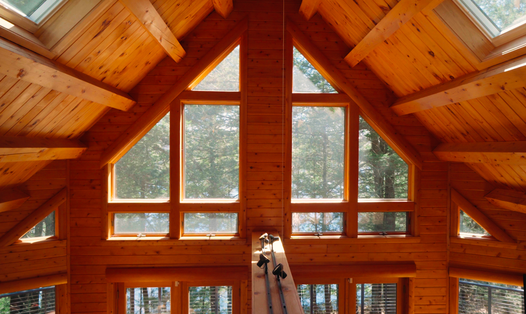 Interior view of a wooden house's attic with large triangular windows showcasing trees outside, exposed wooden beams on the ceiling, and a ladder leaning against the wall.