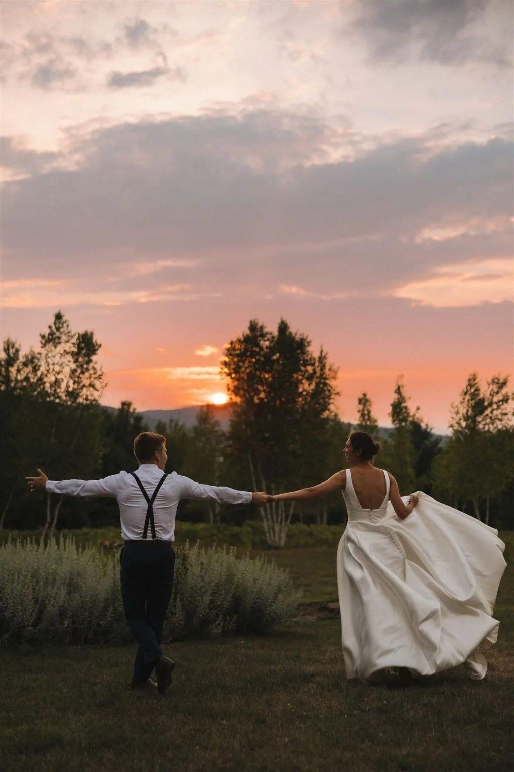 A bride and groom dancing outdoors at sunset, holding hands, with the bride in a flowing white wedding dress and the groom in a white shirt with suspenders.