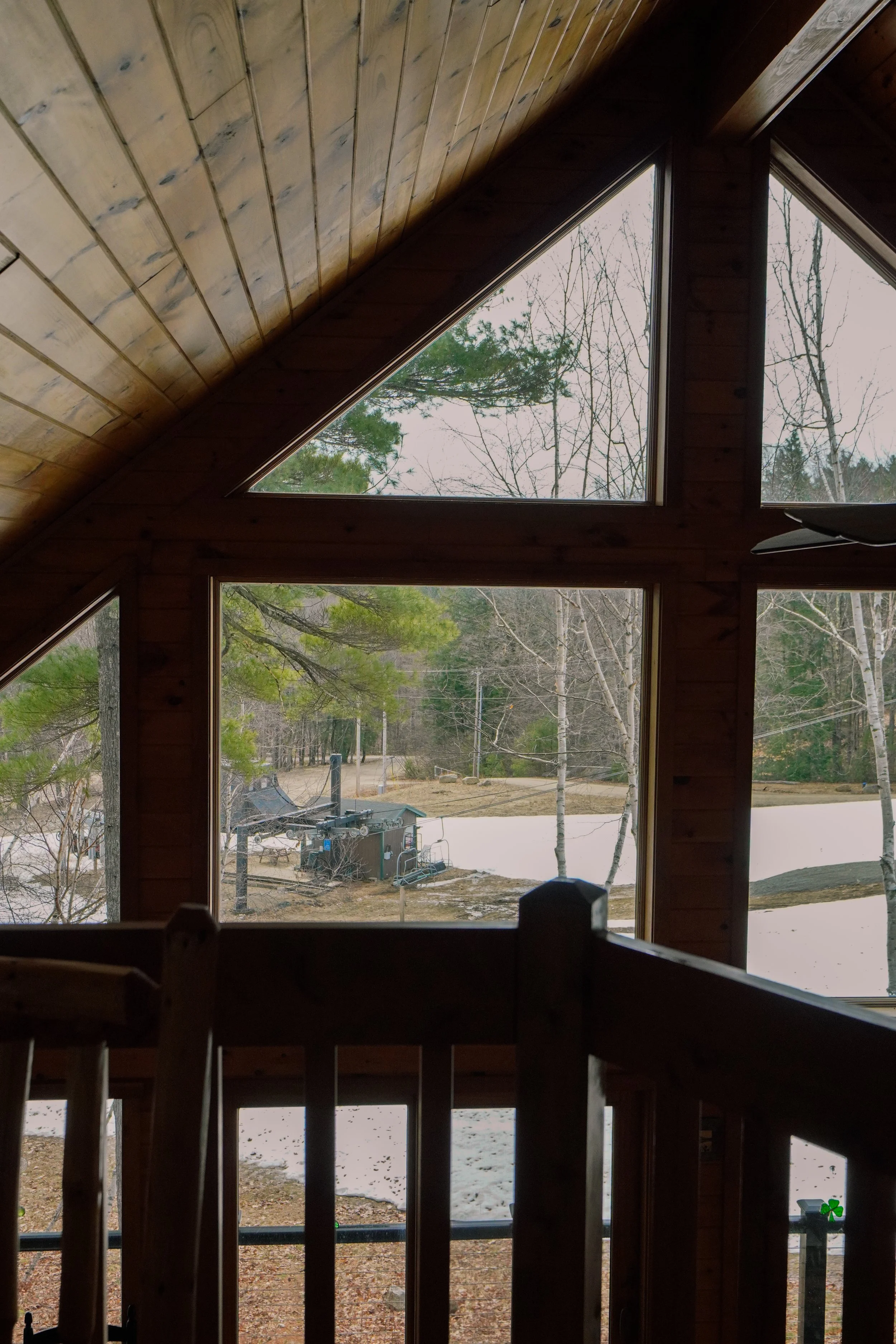 View from inside a house looking out through large, triangular and rectangular windows in an attic or upper floor with wooden walls and ceiling, showing snow-covered ground, trees, and outdoor equipment.