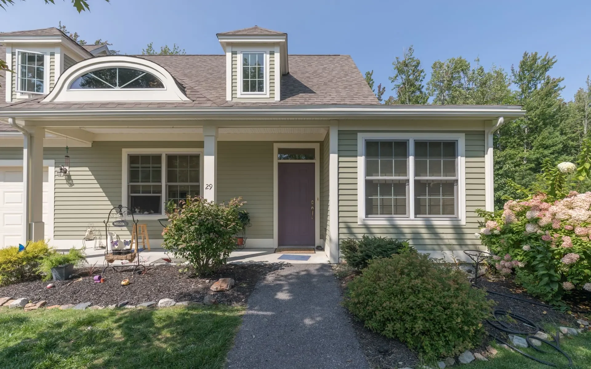 Front view of a beige house with a covered porch, two large windows, and a purple door. The house is surrounded by a garden with bushes and flowers, and a gravel pathway leading to the door.