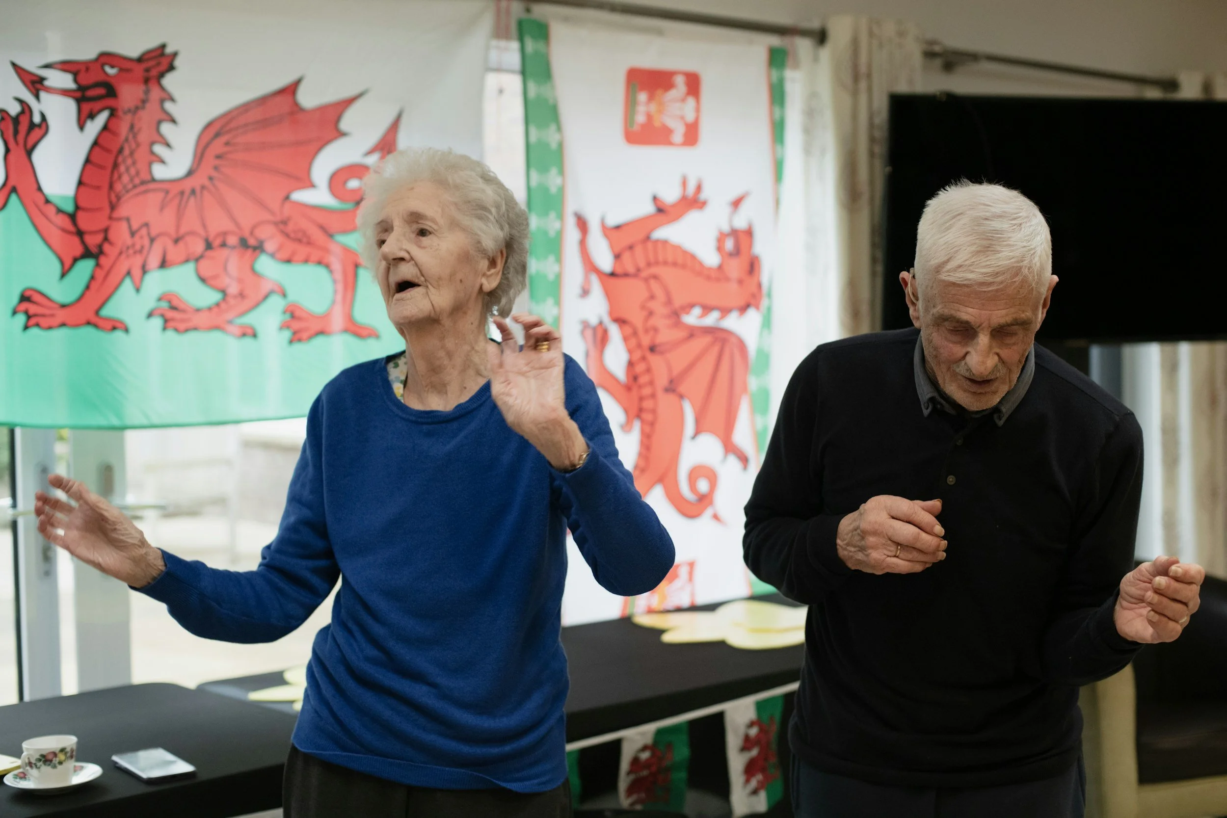 An elderly woman and an elderly man dancing or celebrating indoors, with Welsh flags in the background, featuring red dragons on a green and white banner.