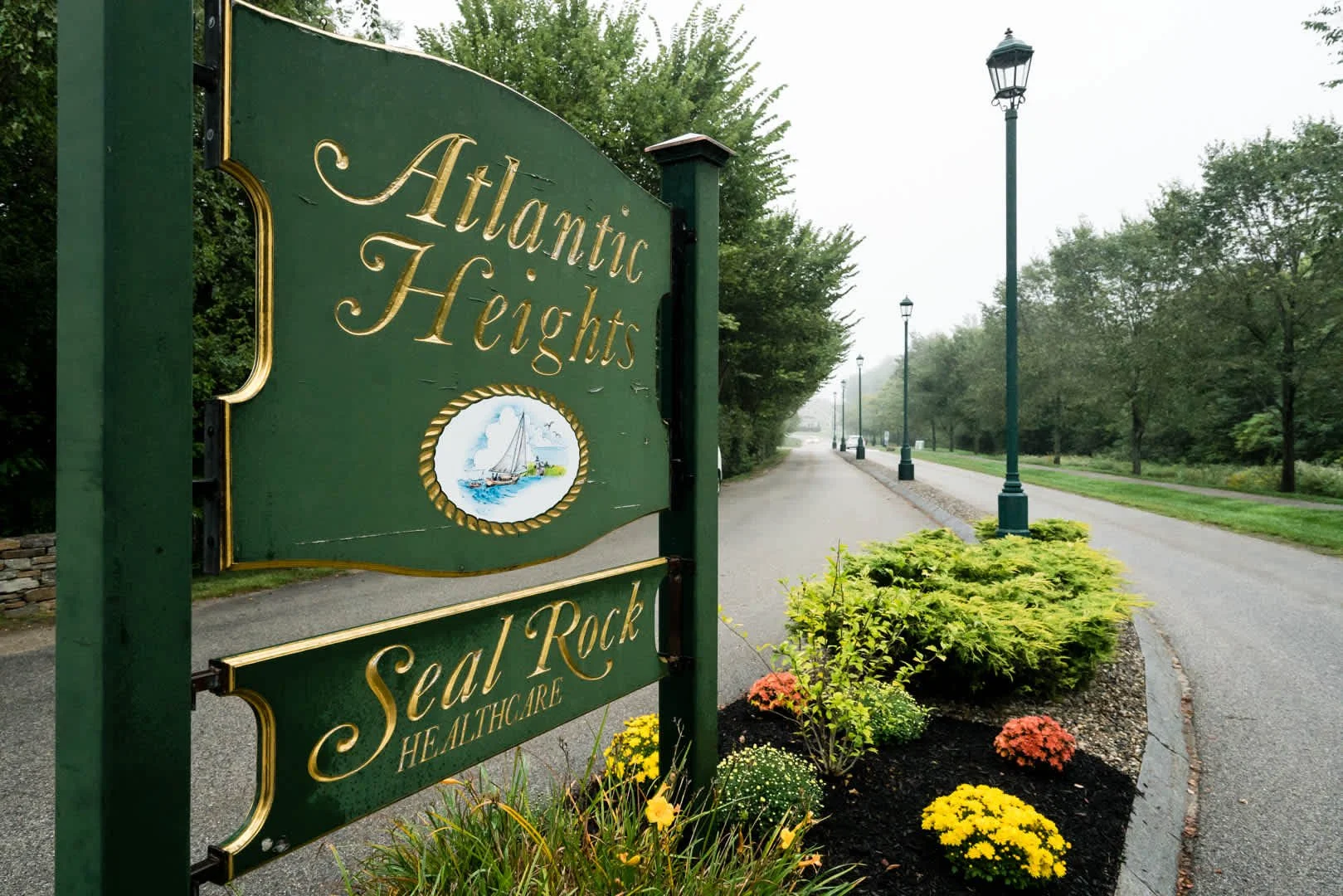 Sign for Atlantic Heights and Seal Rock Healthcare along a paved walkway with park bench, lamp posts, trees, and landscaped flowers.