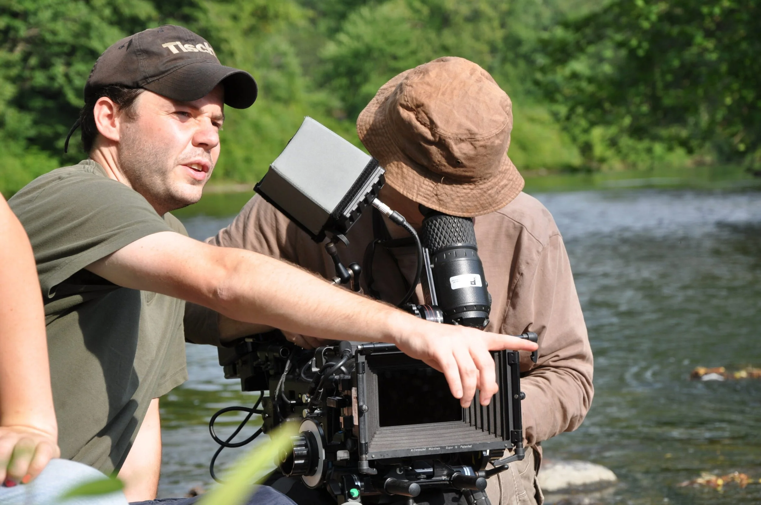 Two men working with a camera on a riverbank surrounded by green trees.