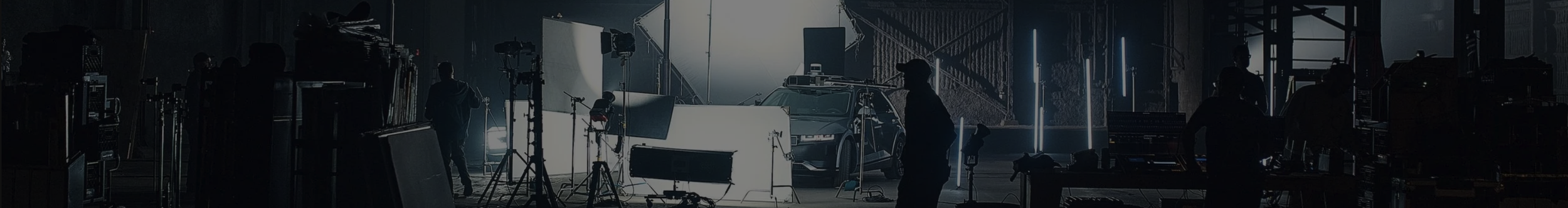 A behind-the-scenes shot of a film or TV production set with crew members, camera equipment, lighting, and a car in a dark studio environment.
