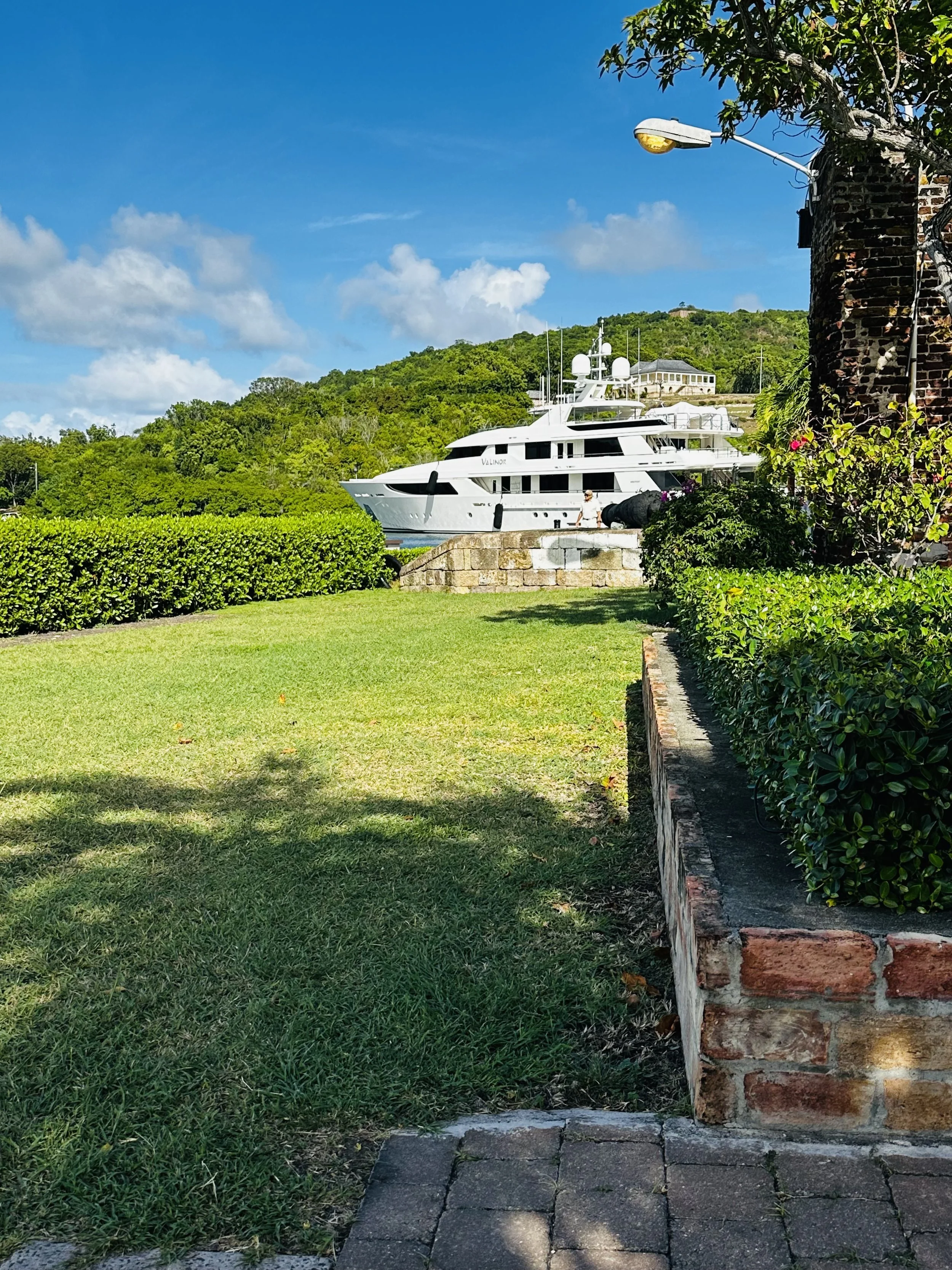 Yacht docked near lush green hill with blue sky and white clouds in the background. Foreground has a grassy lawn, shrubs, and brick walkway.