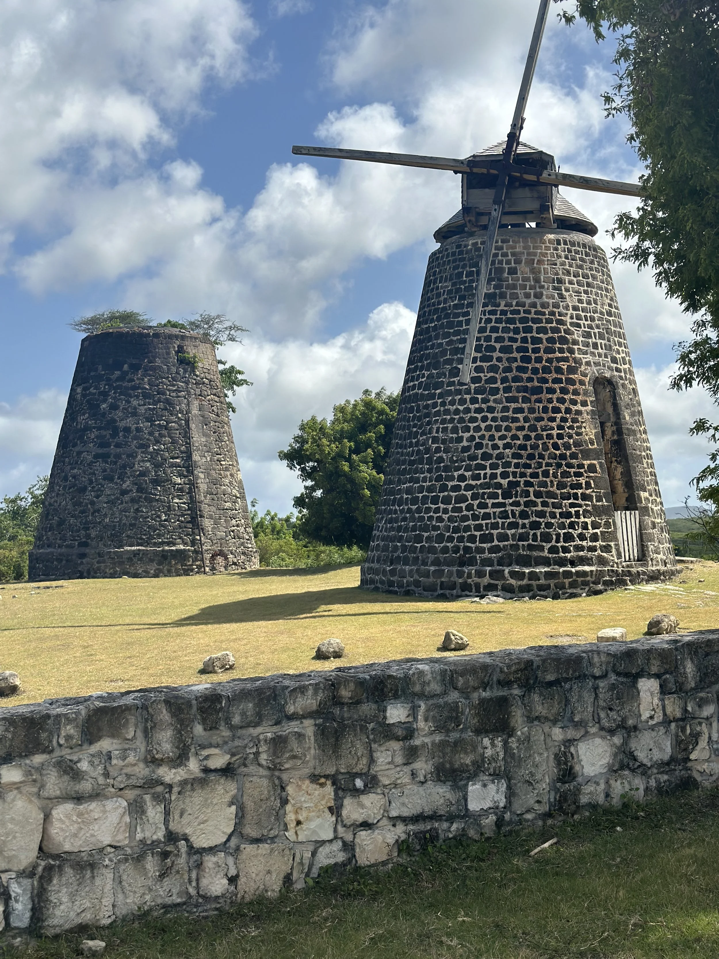 Two old stone windmills, one with blades, in a grassy area with a stone wall in the foreground and trees in the background under a partly cloudy sky.