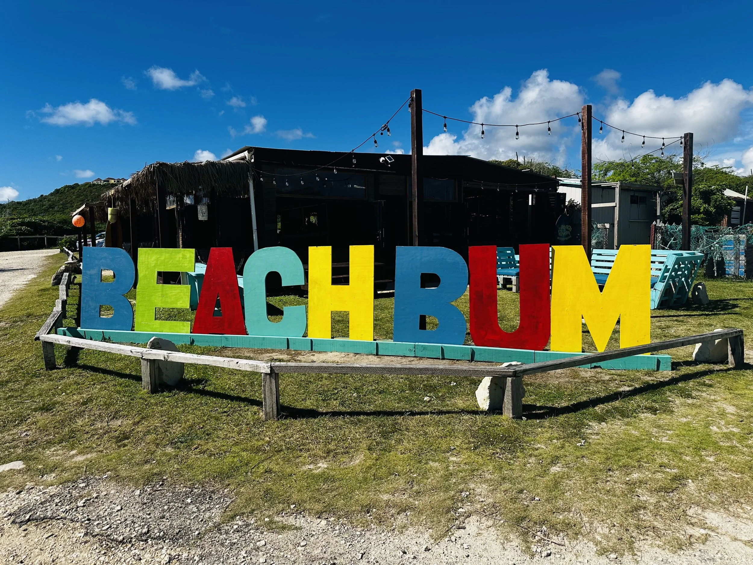 Colorful large wooden letters spelling 'BEACHBUM' are arranged on a grassy area with a wooden border. In the background, there is a dark building with string lights, a few blue chairs, and greenery under a bright blue sky with some clouds.
