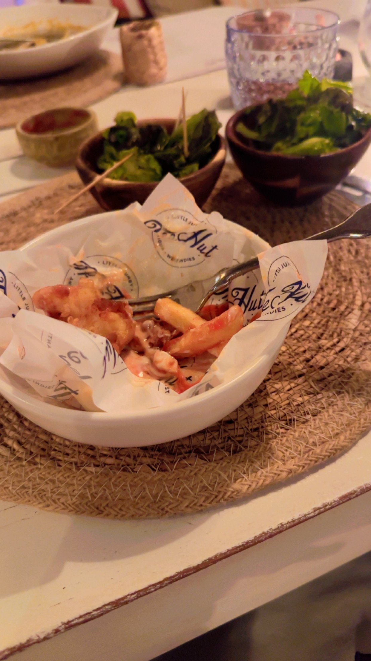 A white bowl with fried shrimp and a fork on a woven placemat on a dining table. In the background, there are two bowls of salad, a glass of water, and other dishes.