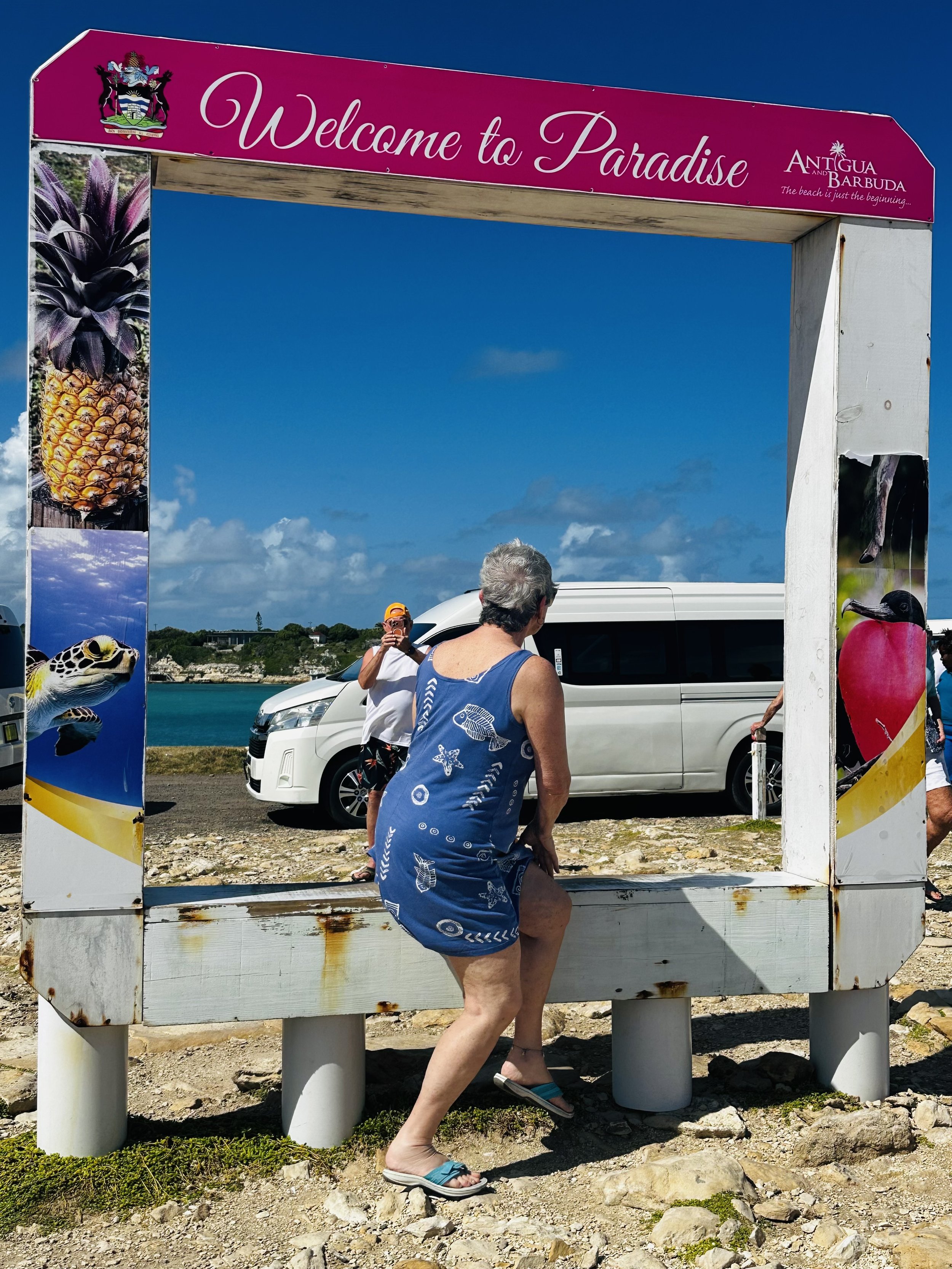 An elderly woman in a blue dress climbing into a large wooden frame with a pink sign that says 'Welcome to Paradise' and images of tropical fruits and animals, by the beach with a parking lot and water in the background.