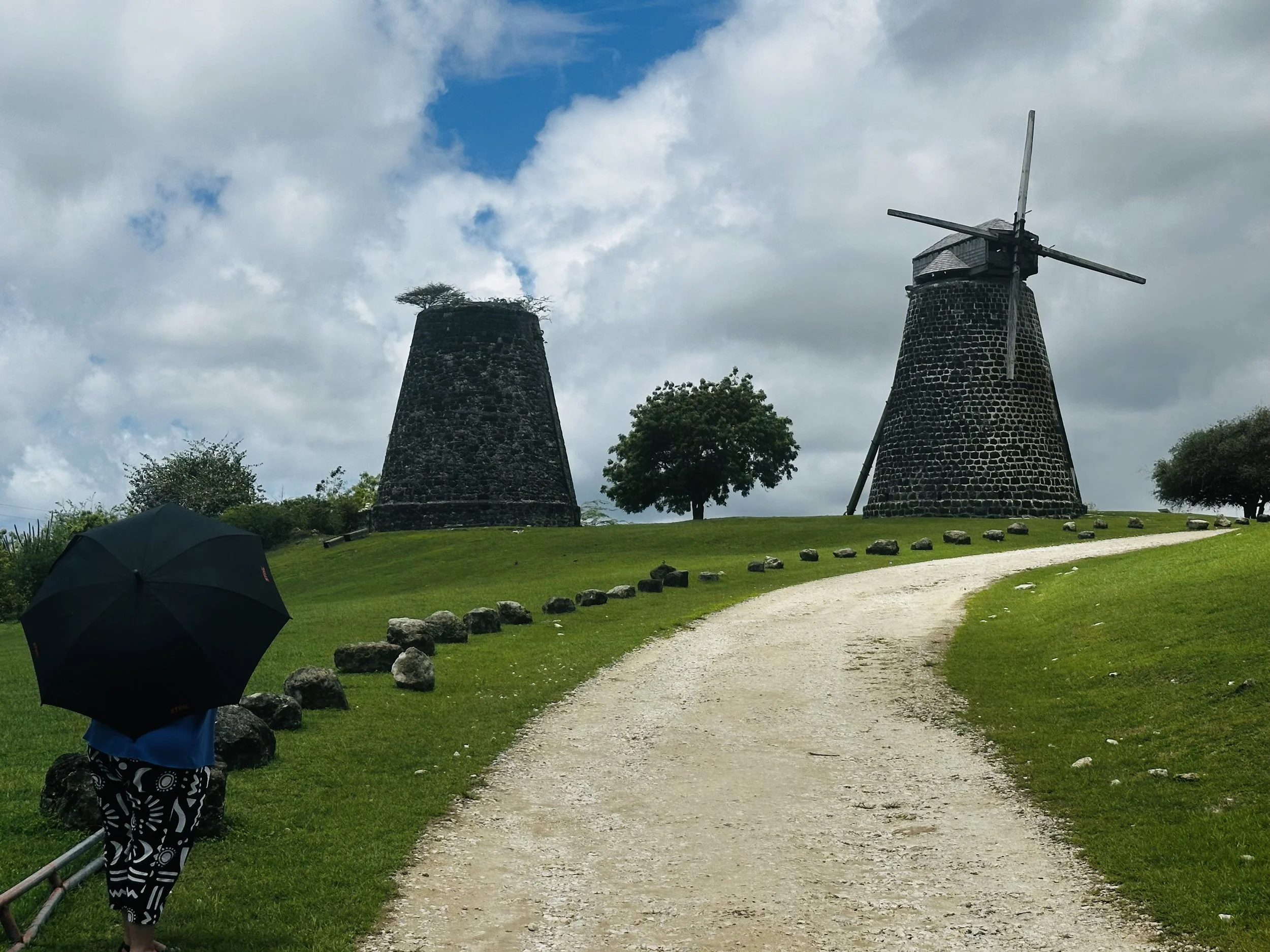 A person with a black umbrella walking along a gravel path on a grassy hill, with two historic stone windmills in the background and a partly cloudy sky.
