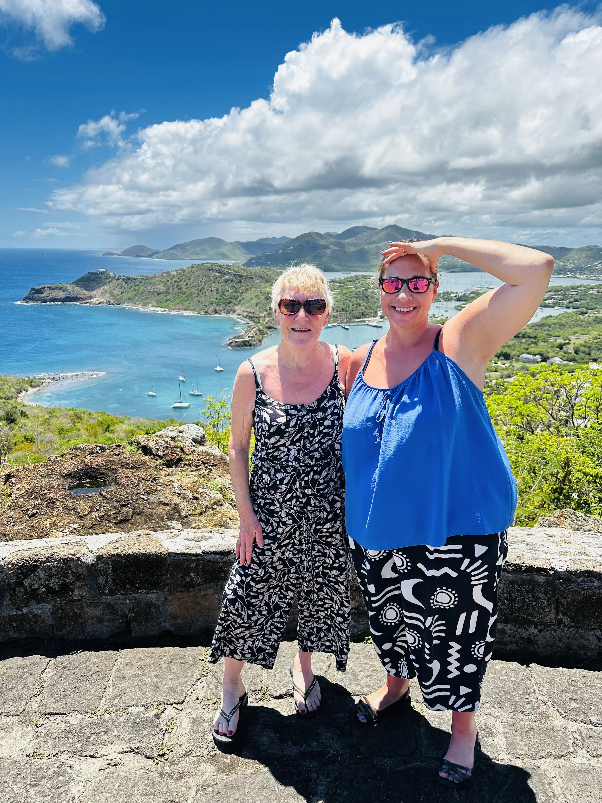 Two women standing on a stone ledge with a scenic view of a coastal bay, green hills, and boats in the water behind them, under a partly cloudy sky.
