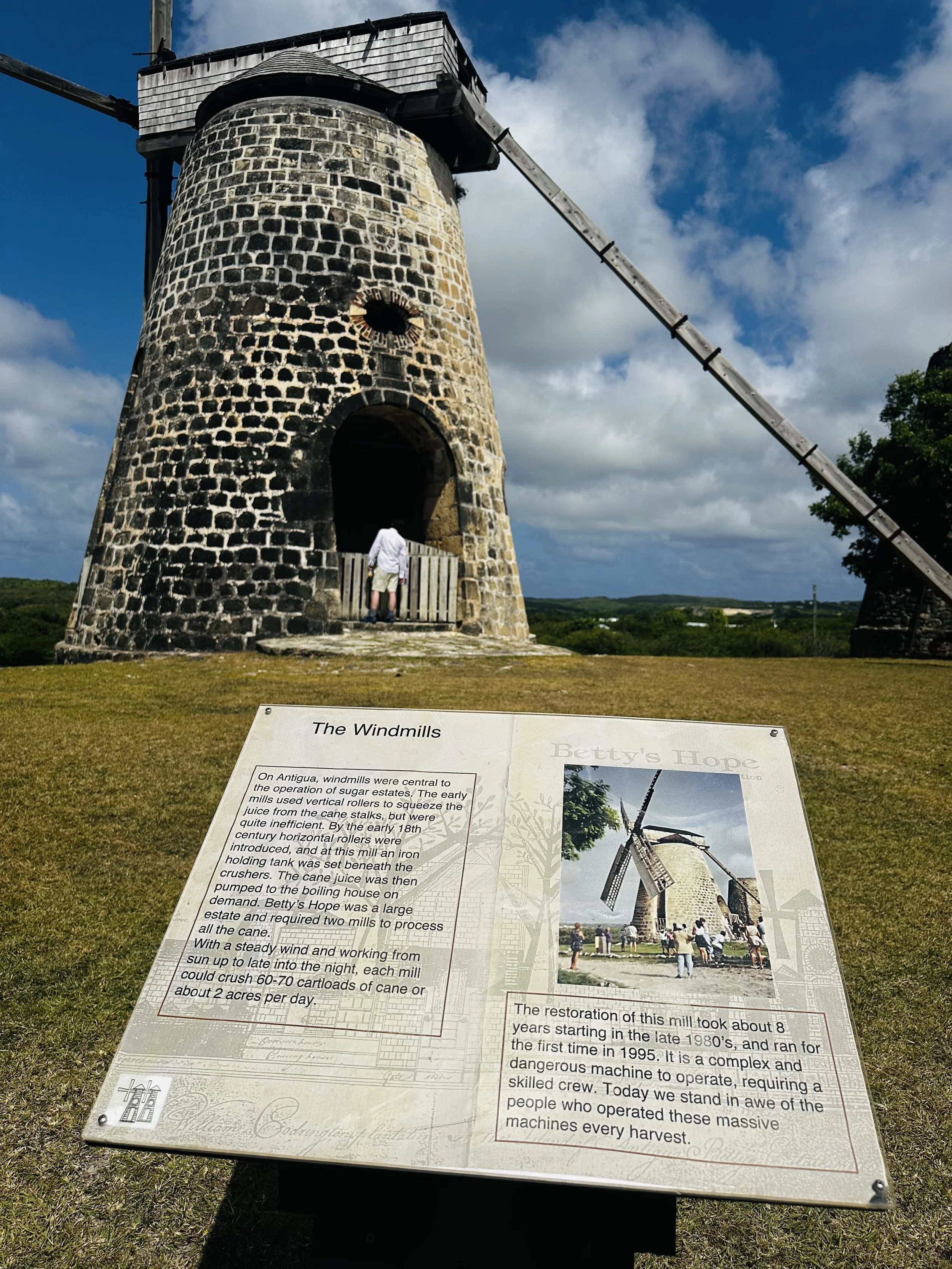 A historic stone windmill with four sails, situated on a grassy landscape under a partly cloudy sky. An informational sign about the windmill is in the foreground. A person stands at the entrance of the windmill.