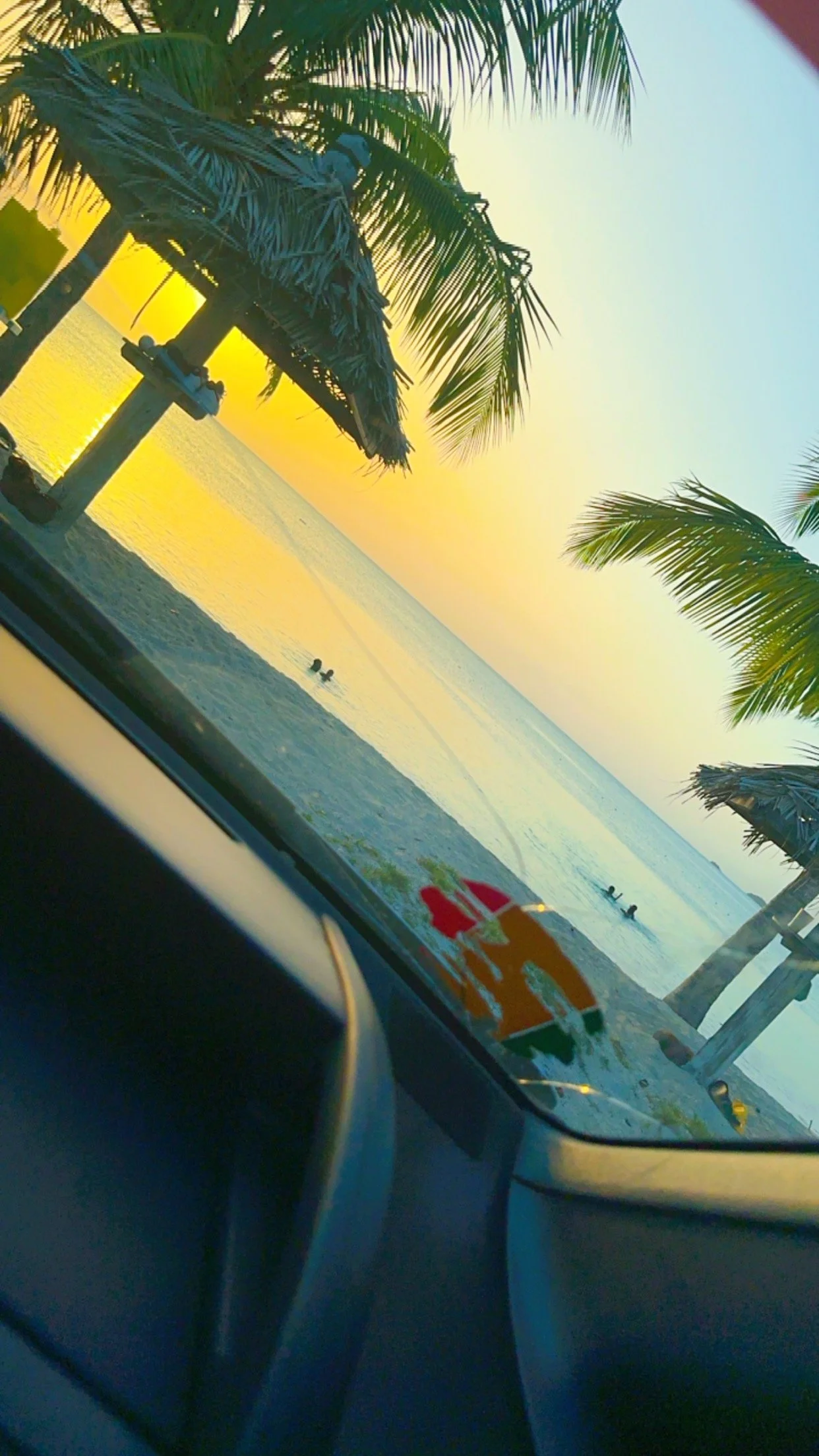 Sunset view over a beach with palm trees and thatched umbrellas, seen from inside a car.
