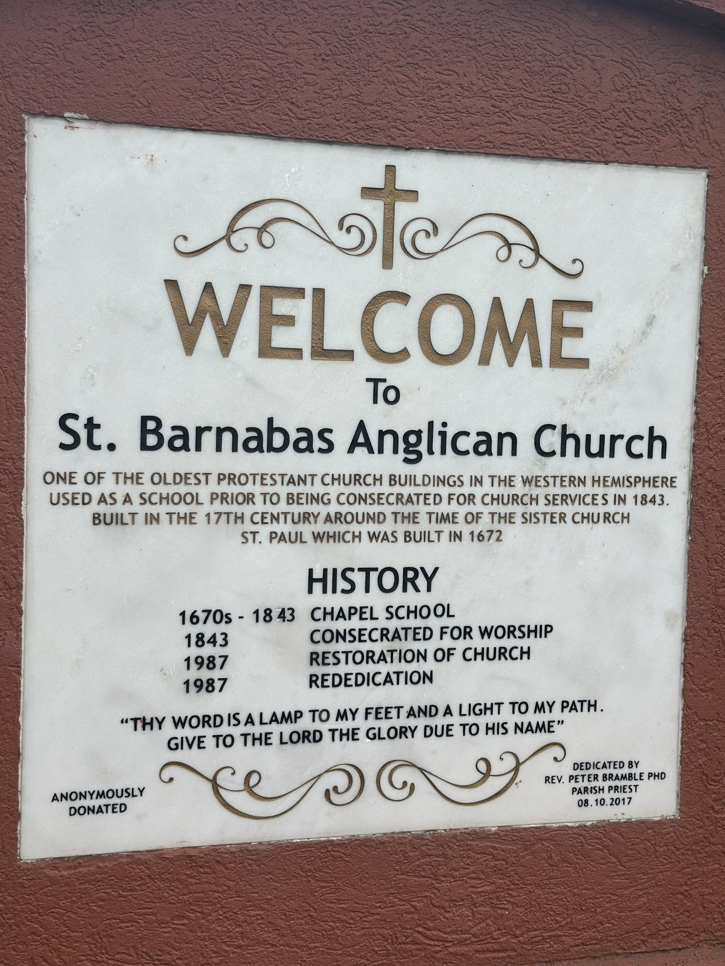 A welcome sign for St. Barnabas Anglican Church with a cross and decorative lines at the top. The sign provides historical information about the church, including its origins as a school, its dedication for worship, and restoration dates. It also fea