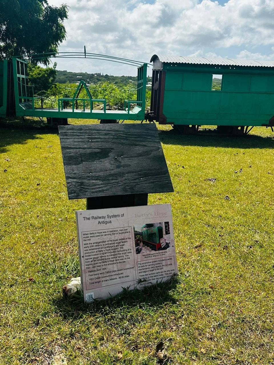 An outdoor display showing a small green model train and a green train car with a curved roof, situated on a grassy area with a sign explaining the railway system of Antigua.