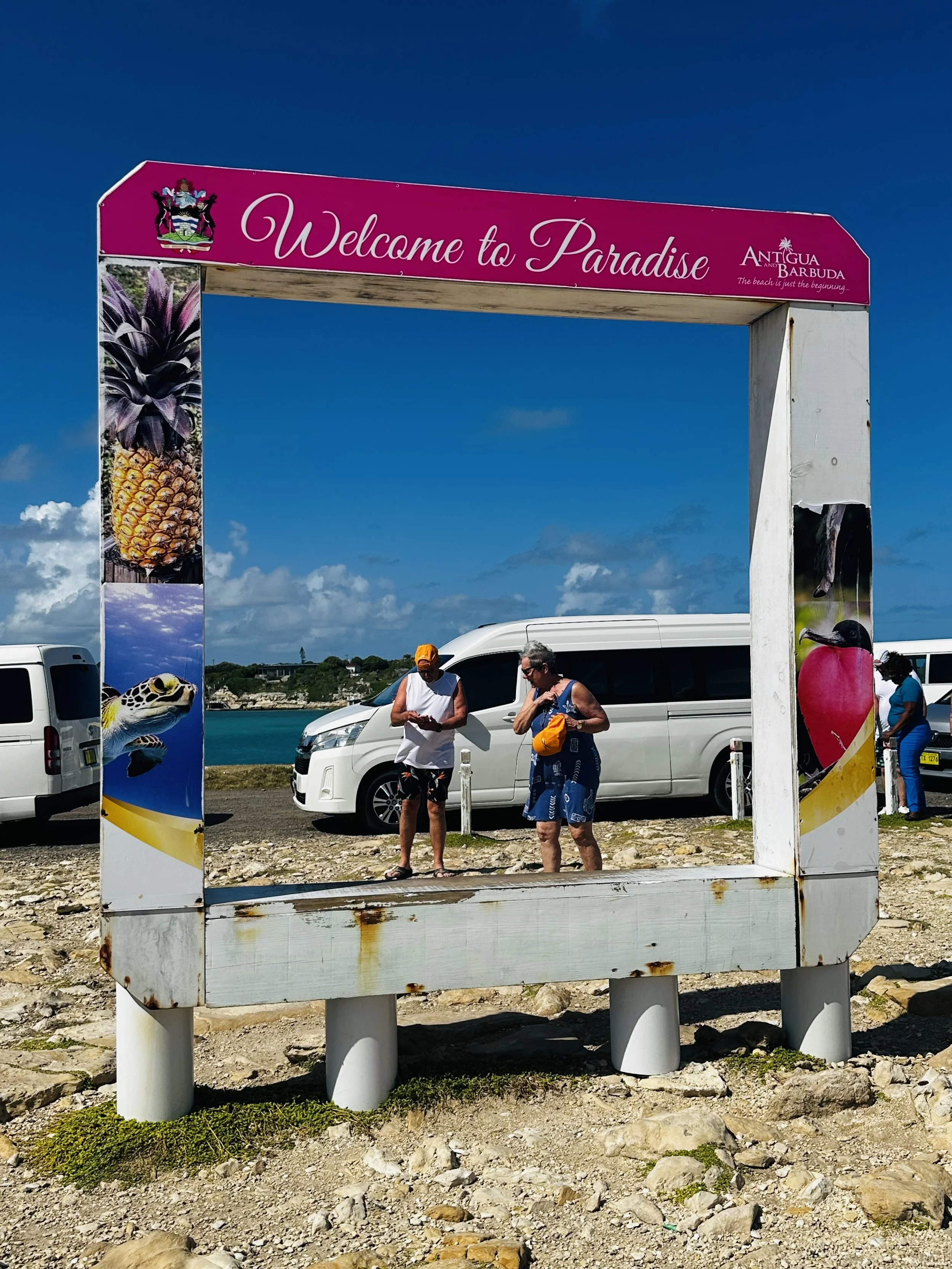 Two women standing in front of a white minivan near the water, framed by a pink and white welcome sign that says "Welcome to Paradise." The sign features images of a pineapple, a turtle, a strawberry, and a beach scene, indicating a tropical location