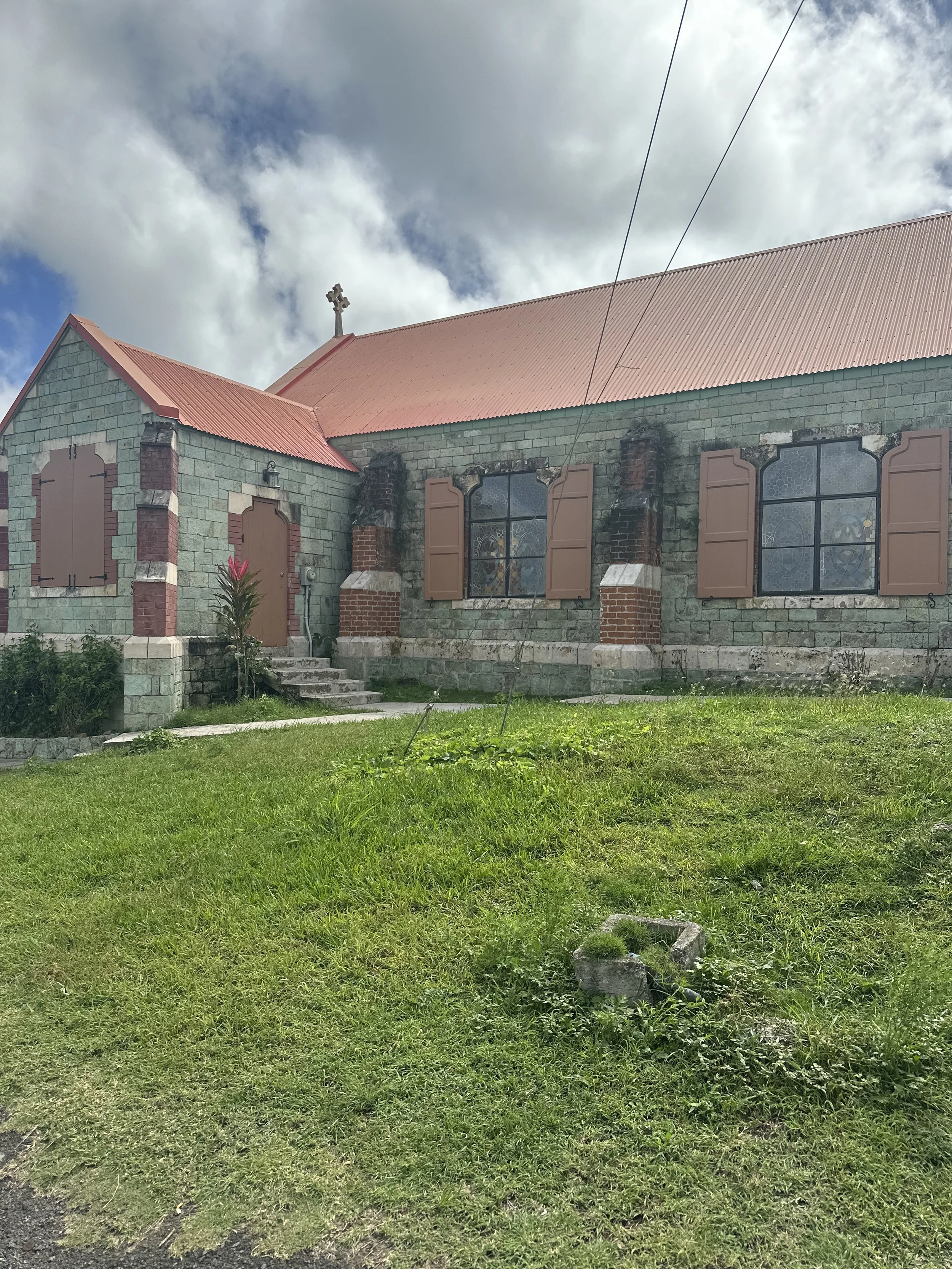 A small stone church with a pink metal roof, stained glass windows, and closed brown shutters on the windows. The church is surrounded by a grassy area with a small flower bush near the steps.