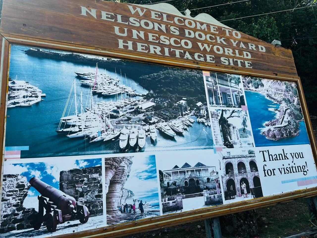Large informational sign welcoming visitors to Nelson's Dock Yard UNESCO World Heritage Site, featuring photos of boats in the harbor, historic cannons, a beach scene, and colonial buildings.