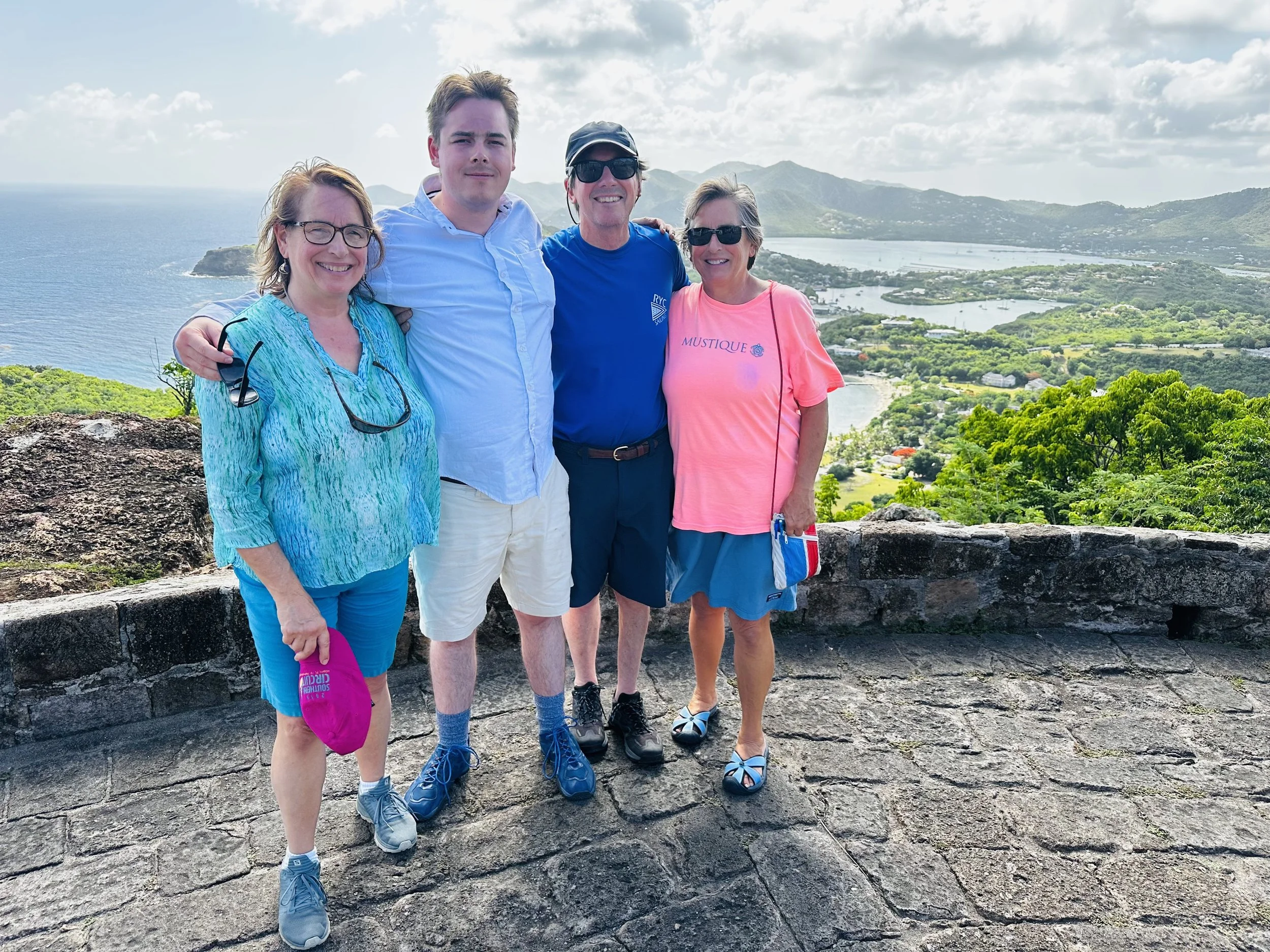 Four people standing on a stone walkway with a scenic view of water, mountains, and green landscape behind them.