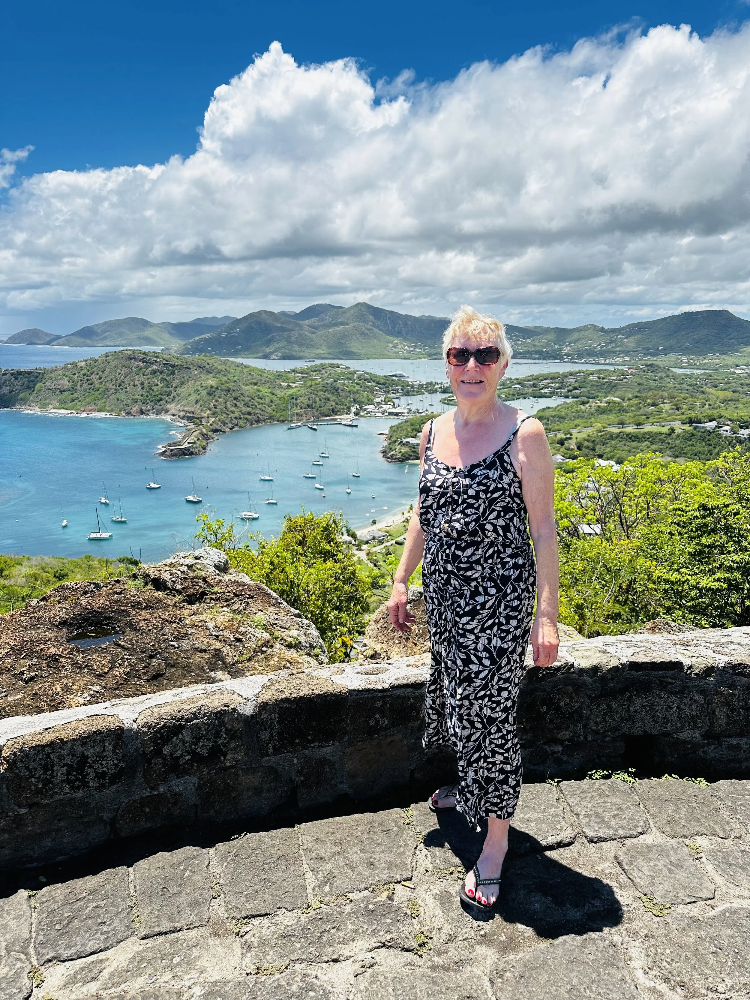 A woman stands on a stone pathway overlooking a tropical bay with sailboats, lush green islands, and mountains in the background, under a partly cloudy sky.