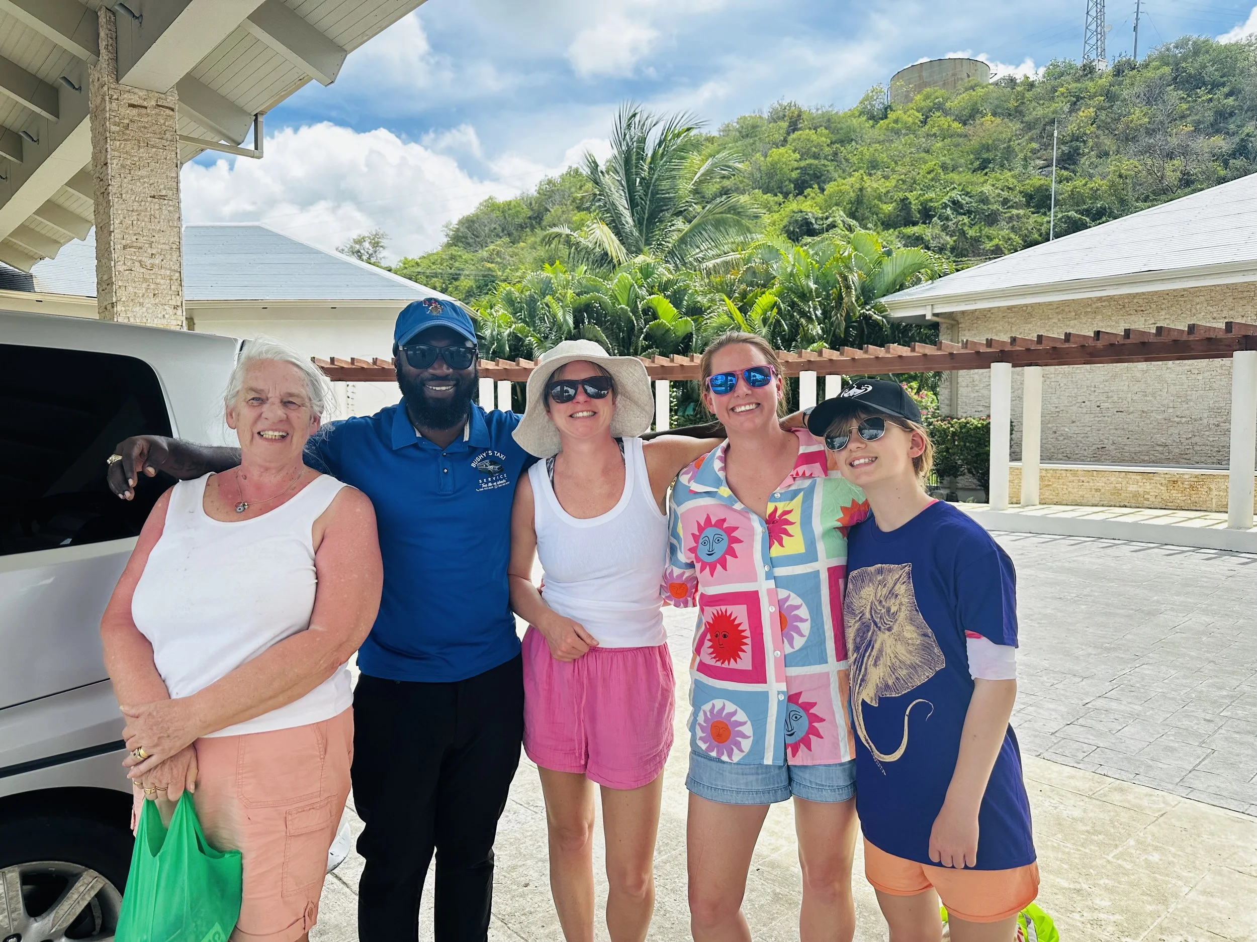Group of five smiling people standing outdoors near white building, palm trees, and green hills, dressed in summer clothes.